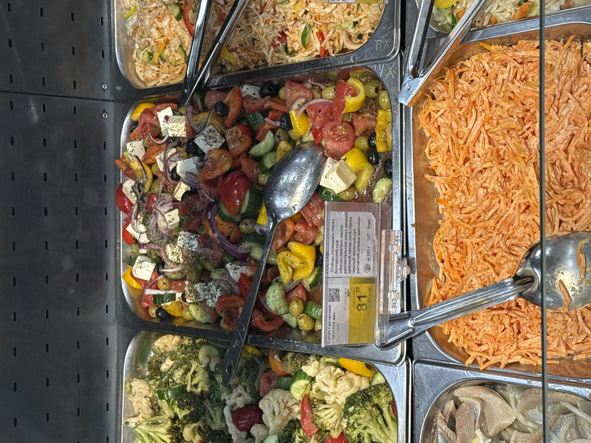 The image shows a refrigerated deli case with several bins of prepared salads. Clockwise from the top left: coleslaw-type salad, Greek salad with feta, shredded carrot salad, pickled herring. Broccoli and cauliflower salad is in the bottom left corner. There are serving utensils present. There is a price tag for the Greek salad, indicating it is from Russia in Izhevsk, and the price is 81.59.