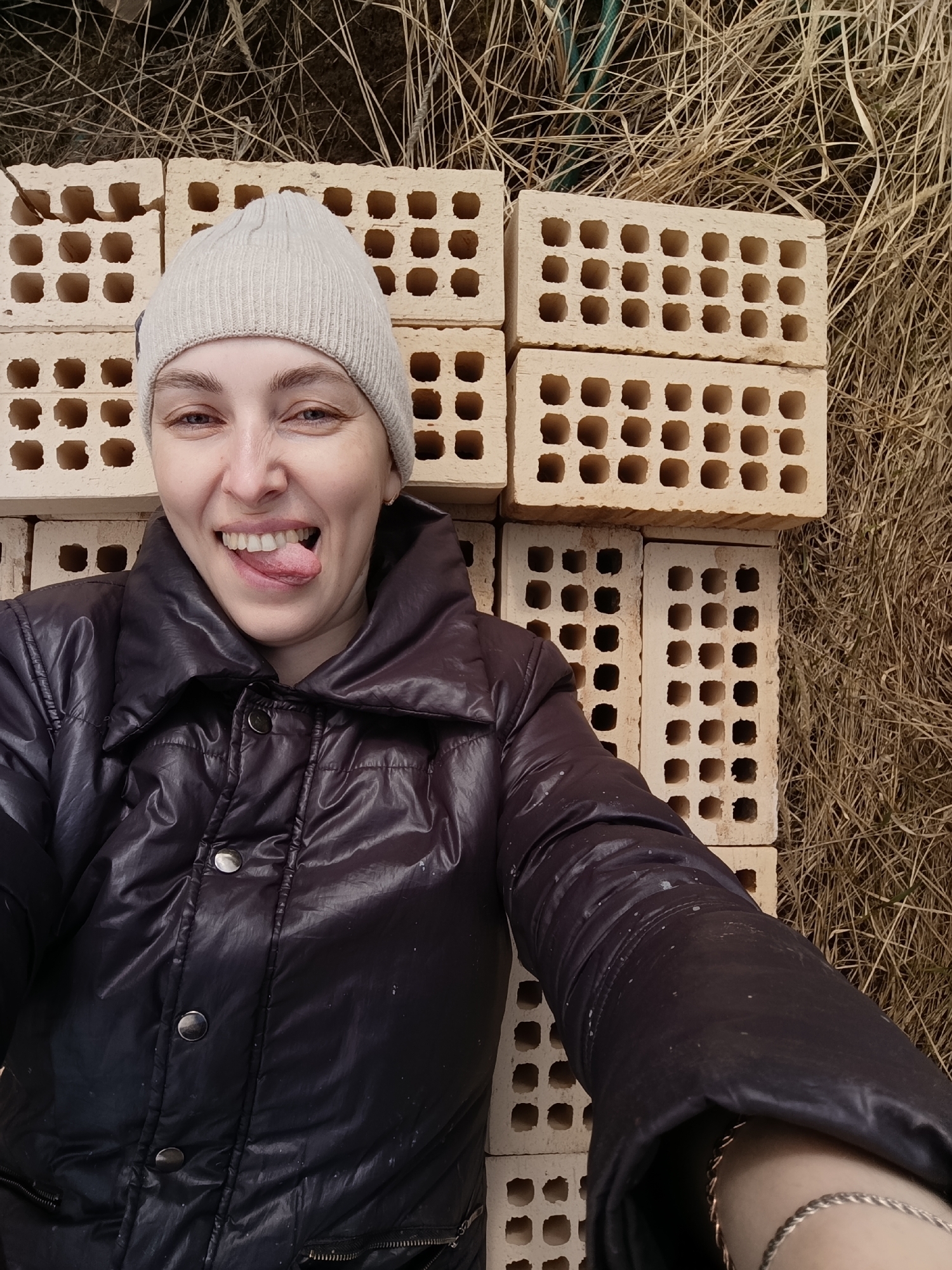 A woman wearing a light-colored beanie and a black puffer jacket lies on her back on a surface consisting of stacks of light-colored bricks and dry, brown grass. Her head rests on the bricks. She is sticking her tongue out and smiling playfully. The location is outside, with natural light, likely in the city of Chaikovsky, Russia.