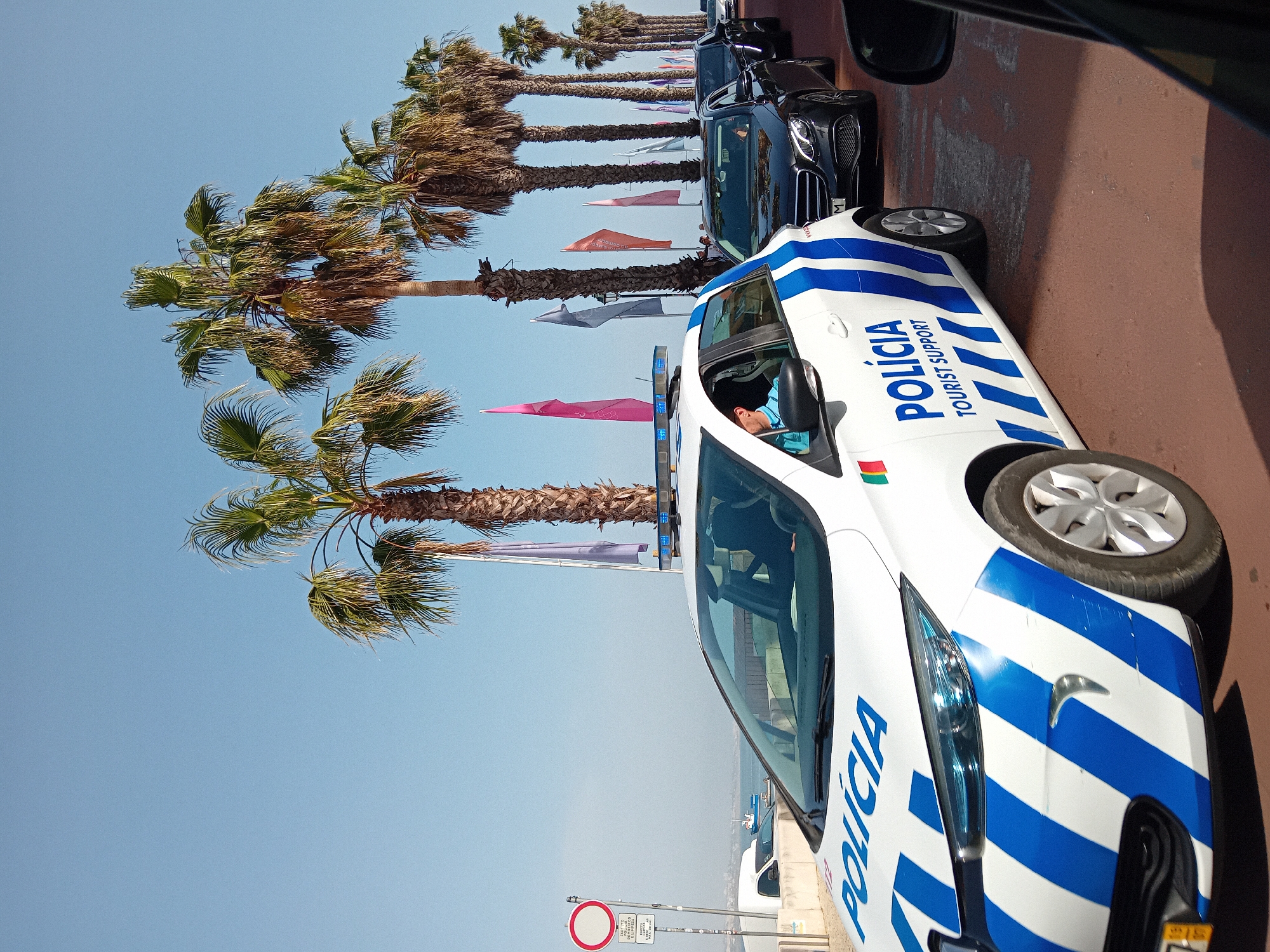 A brightly lit outdoor shot features a white and blue "POLÍCIA TOURIST SUPPORT" car, likely a police vehicle in Cascais, Portugal, parked on a sidewalk or a roadway. The car is situated parallel to a row of tall palm trees, some adorned with colorful flags that seem to represent various countries. Other vehicles are parked behind the police car. The sky is clear and blue. The police car is angled towards the viewer and occupies a substantial portion of the foreground. A person is visible inside the car. A traffic sign stands next to the car. The setting suggests a coastal area, possibly near a beach or tourist-frequented zone in Cascais.