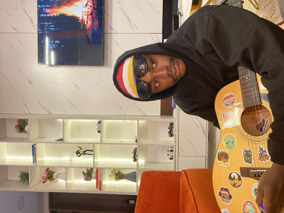 A man wearing a black hoodie, sunglasses, and a colorful beanie is holding a guitar covered in stickers. He is indoors, possibly in a living room. Behind him is a white shelf with decorations and a TV showing a fire. The couch nearby is orange. The photo is taken at an angle, giving the impression that the man is leaning towards the camera.