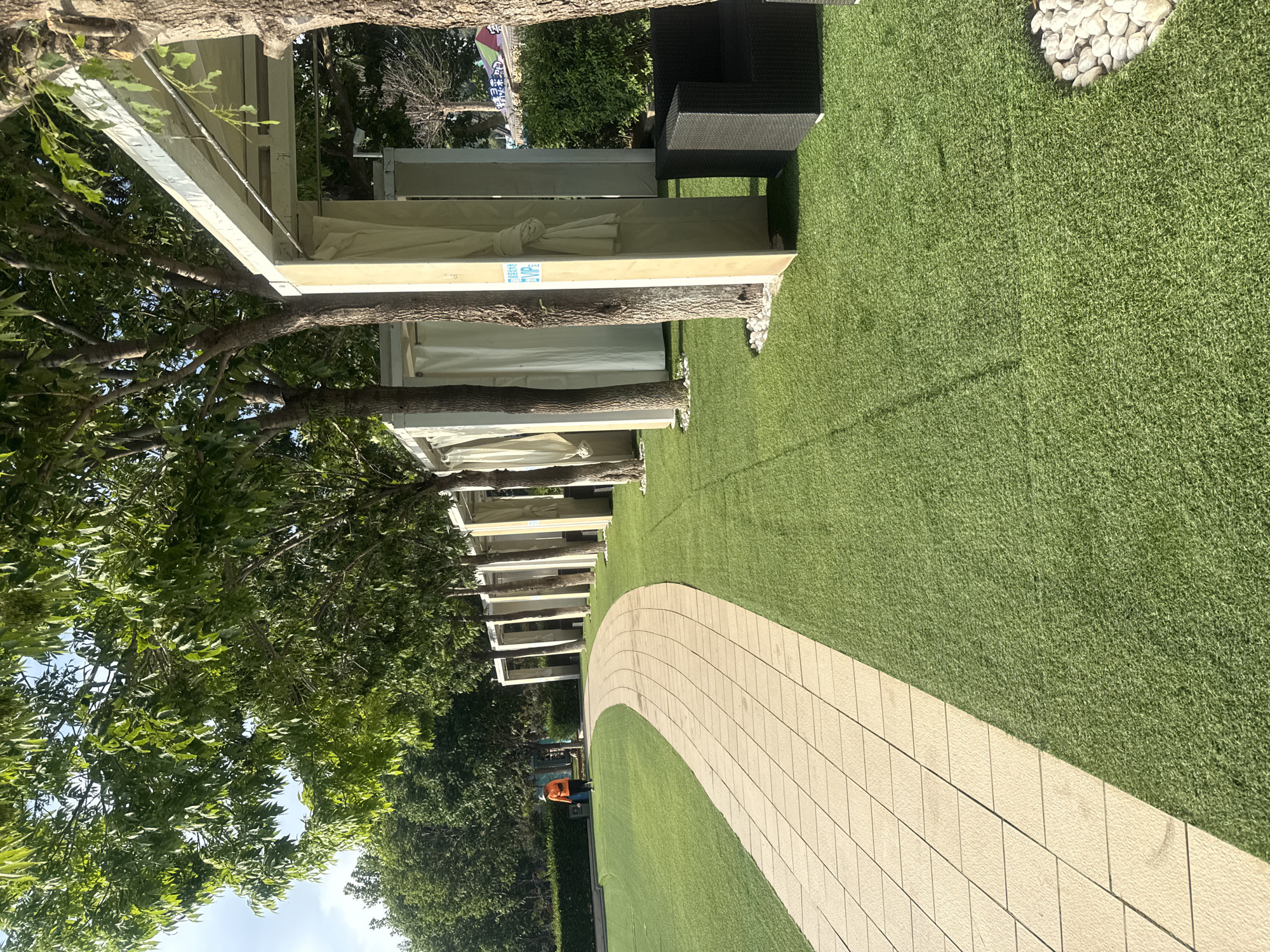 A person in an orange vest and a white hat is walking away from the camera on a paved pathway. The pathway curves to the left and is bordered by a lawn of artificial turf. To the right of the pathway are a row of white cabanas with beige curtains, each supported by light-colored pillars. Trees with green foliage grow between the cabanas and along the pathway. A dark-colored armchair is visible next to one of the cabanas. The scene is set in an outdoor park-like area. This image was taken in the city of Qinhuangdao, China.
