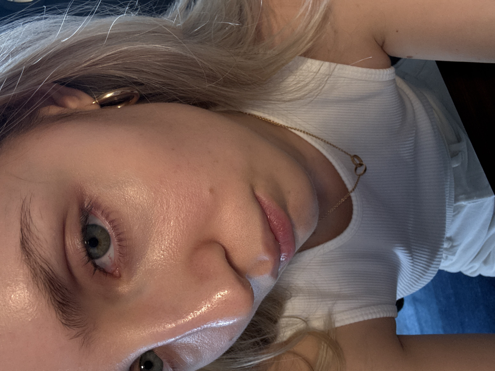 A close-up, tilted perspective photograph captures a person with fair skin, blonde hair, and green eyes. They wear a white ribbed tank top and a gold hoop earring. A delicate gold necklace rests against their skin. The environment suggests an interior setting, with dark and blue tones visible in the background, potentially from clothing or furnishings. The location within Mexico City, Mexico, cannot be determined from the visual information provided.