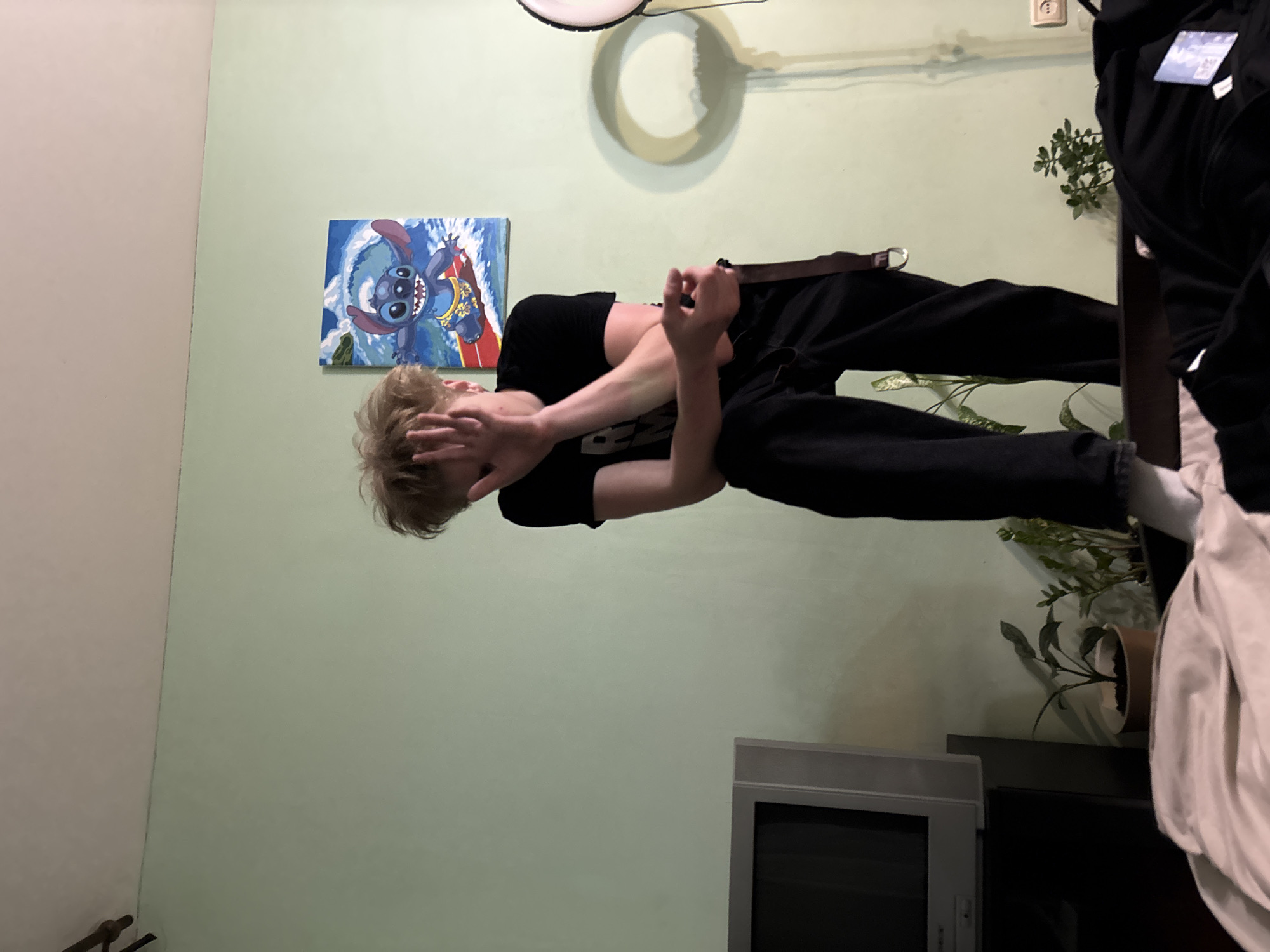 In an indoor environment, a young person with blonde, tousled hair is captured in a moment of apparent distress or shyness, obscuring their face with their right hand. They are dressed in dark clothing, including a black t-shirt and black pants with suspenders. The individual is positioned in a relaxed yet somewhat contorted pose, leaning against an unseen surface with their right elbow resting on their knee.
The background of the scene is dominated by a light green wall. On this wall, a colorful painting of the cartoon character Stitch surfing on a red surfboard amidst a blue wave is displayed. To the left, a vintage television set sits on a stand. Several potted plants are visible, adding a touch of greenery to the room. A circular shadow, possibly cast by a light fixture, is prominent on the wall to the right.
The floor area in the foreground is partially covered by a light-colored duvet or blanket, and a dark garment, possibly a jacket, lies folded on top. The overall impression is of a personal space, likely a bedroom or living area, within a dwelling. The specific location in "Unknown, Russia" is not discernible from the visual cues present in the image.