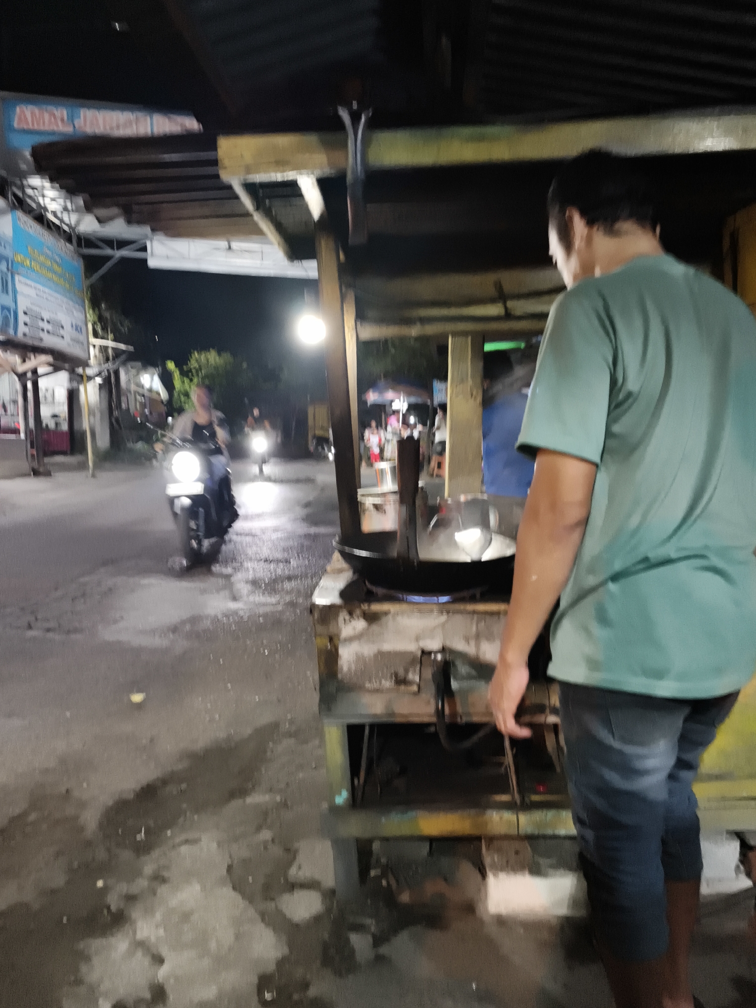 The image captures a nighttime street scene in Tempel, Indonesia. In the foreground, a person wearing a teal t-shirt and denim shorts stands by a food cart, looking down at a large wok placed on a stove. The wok appears to be cooking something, with steam or smoke rising from it. The food cart itself is rustic, made of wood and metal.
In the background, the street is wet, suggesting recent rain or damp conditions, and reflects the light from various sources. Two motorcycles are visible on the street. The one closer to the viewer is ridden by a person wearing a dark outfit and a light-colored shawl or jacket. Their face is blurred due to motion. The second motorcycle further down the street is also illuminated by its headlights.
Several signs are visible along the street. One sign, partially obscured, has blue and red text and appears to be an advertisement or public announcement. Another sign, to the left, is predominantly blue and white with text in Indonesian. The overall lighting suggests it is evening or night, with artificial lights illuminating the area. The atmosphere seems to be that of a bustling local street, possibly a marketplace or a roadside food stall area.