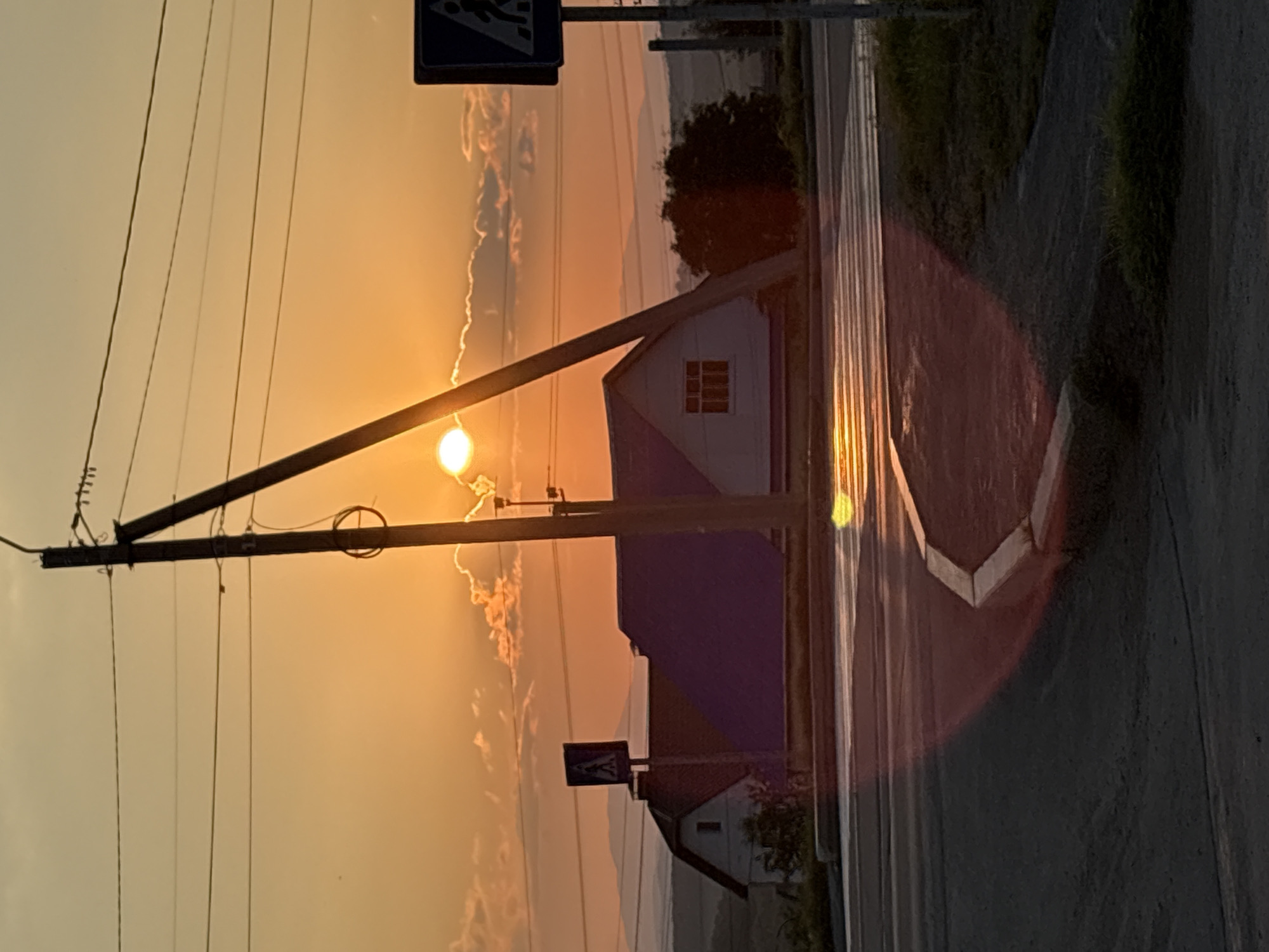 A low-angle, vertical, medium shot captures a rural road at dusk, illuminated by a hazy orange sunset that peeks from behind scattered clouds. The sun, a bright, diffused orb, casts a warm glow across the scene. In the foreground, a paved road curves to the left, with a concrete curb on the right side. The road appears wet, reflecting the sunset's light.
Behind the road, a house with a dark, angled roof and white walls is visible. A pedestrian crossing sign stands on a pole to the left of the house. Two utility poles stand prominently in the middle ground, one diagonally bisecting the frame and supporting multiple wires. Another utility pole is visible on the far right, also supporting wires, and topped with a pedestrian crossing sign. The background features distant, muted mountains and a silhouette of trees. The overall atmosphere is serene and tranquil.