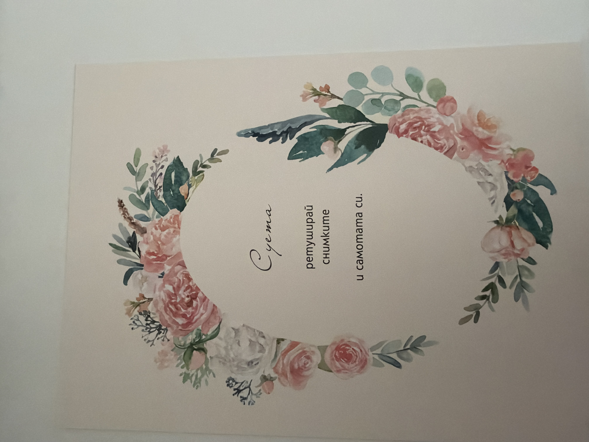 The image is a close-up, high-angle shot of a card with a floral design. The card has a pale, creamy background. A watercolor-style floral wreath, featuring pink and white roses, greenery, and other delicate flowers, encircles an oval-shaped area in the center of the card. Within this oval, there is text written in Bulgarian. The first line of text, in a cursive font, reads "Суета". Below this, in a smaller, sans-serif font, are two lines: "ретуширай" and "снимките". The bottom line of text, in the same sans-serif font, reads "и самотата си.". The floral elements are positioned on the left, right, and top edges of the oval, with some extending slightly beyond the oval's boundary. The lighting is soft and even, suggesting an indoor setting. There is no information in the image to indicate a specific location in the city of Pazardzhik, Bulgaria.