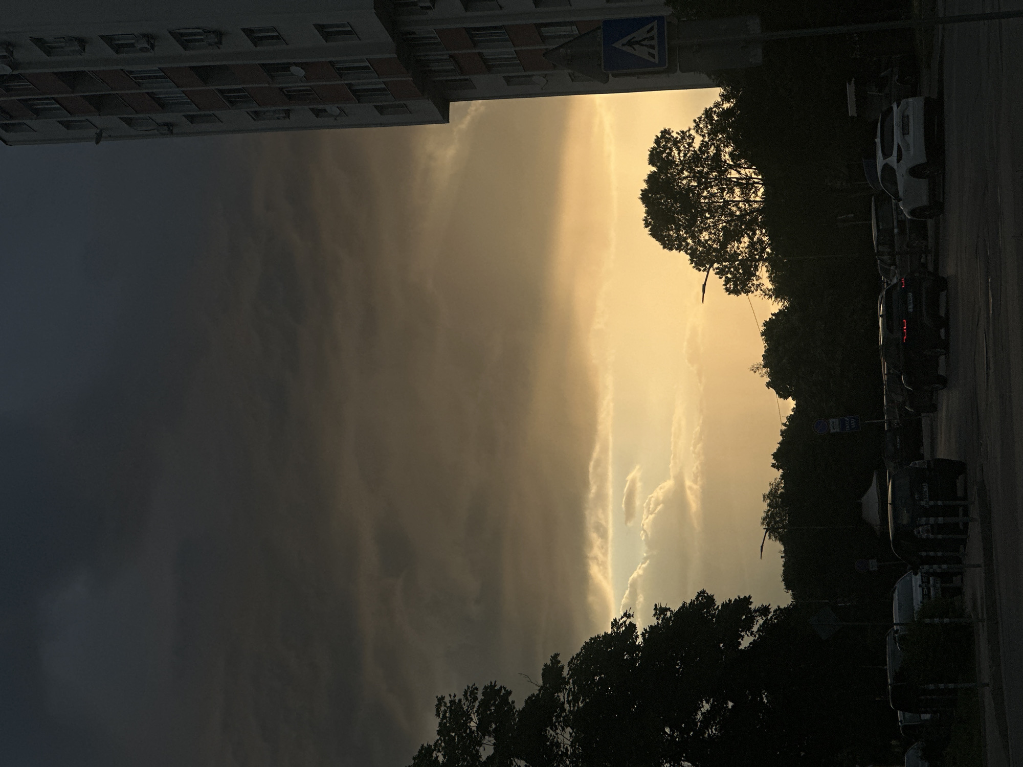 This is a photograph of a street scene in Sofia, Bulgaria, taken at dusk or during stormy weather, as indicated by the dark, dramatic clouds in the sky and the low light conditions. The image captures a section of a city street with several cars parked or driving, silhouetted against the muted glow of the sky.
On the right side of the frame, a tall, multi-story building with a light-colored facade and red accents dominates the upper portion. Numerous windows and what appear to be air conditioning units are visible on its side. Adjacent to the building, a blue pedestrian crossing sign is mounted on a pole.
The sky is filled with heavy, dark clouds that transition to lighter, yellowish hues near the horizon, suggesting either the setting sun or dramatic weather phenomena. Fluffy clouds are visible in the lower sky area, catching the light.
In the foreground and mid-ground, several vehicles are present, including a white SUV, a dark-colored car, and other unidentifiable vehicles. The trees and foliage in the scene are dark silhouettes, adding to the overall somber mood. A street lamp is visible in the distance, partially obscured by trees. The overall impression is one of an urban environment experiencing inclement weather or twilight, with the building and vehicles acting as the primary human-made elements within the natural dramatic sky.