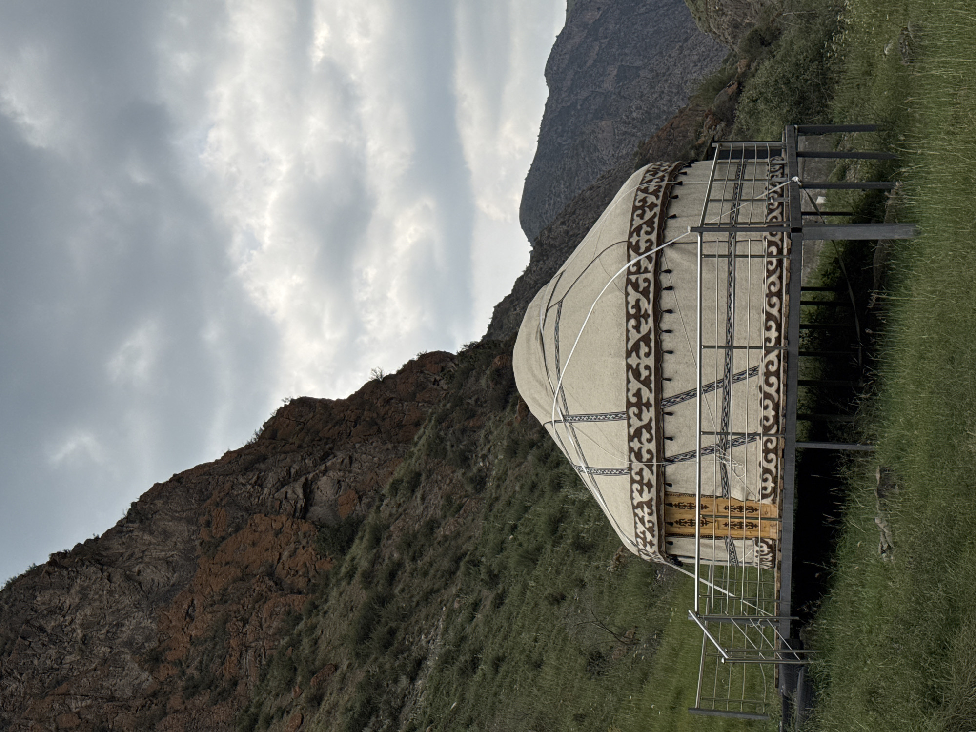The image depicts a traditional Kyrgyz yurt situated on a raised platform in a mountainous landscape. The yurt, made of canvas and supported by a wooden frame, is decorated with patterned trim. A metal railing surrounds the platform, which has stairs leading down to the grassy ground. The surrounding environment consists of steep, rocky slopes covered in sparse vegetation and a cloudy sky overhead. In the background, distant mountains are visible. The scene is serene and appears to be in a natural, undeveloped area. The location is identified as the city of Kyzyl-Korgon, Kyrgyzstan.