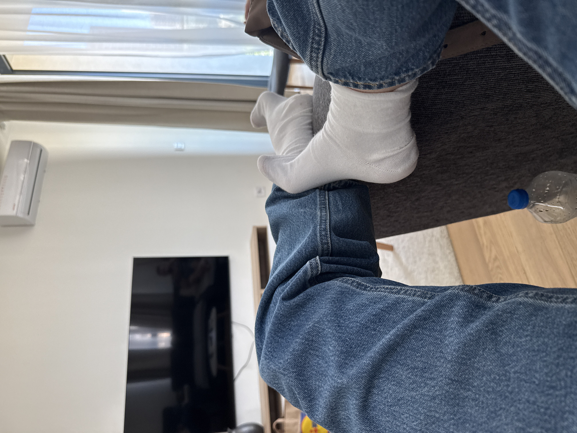 A medium, angled, indoor shot captures a person sitting at a table, wearing blue jeans and white socks, with one foot crossed over the other knee. A bottle of water is on the table next to the person. The interior background includes a window with light-colored curtains, a white wall with a white air conditioning unit, and a black flat-screen television. The location appears to be Belgrade, Serbia, although this cannot be conclusively determined from the image. No people or other actions are visible.