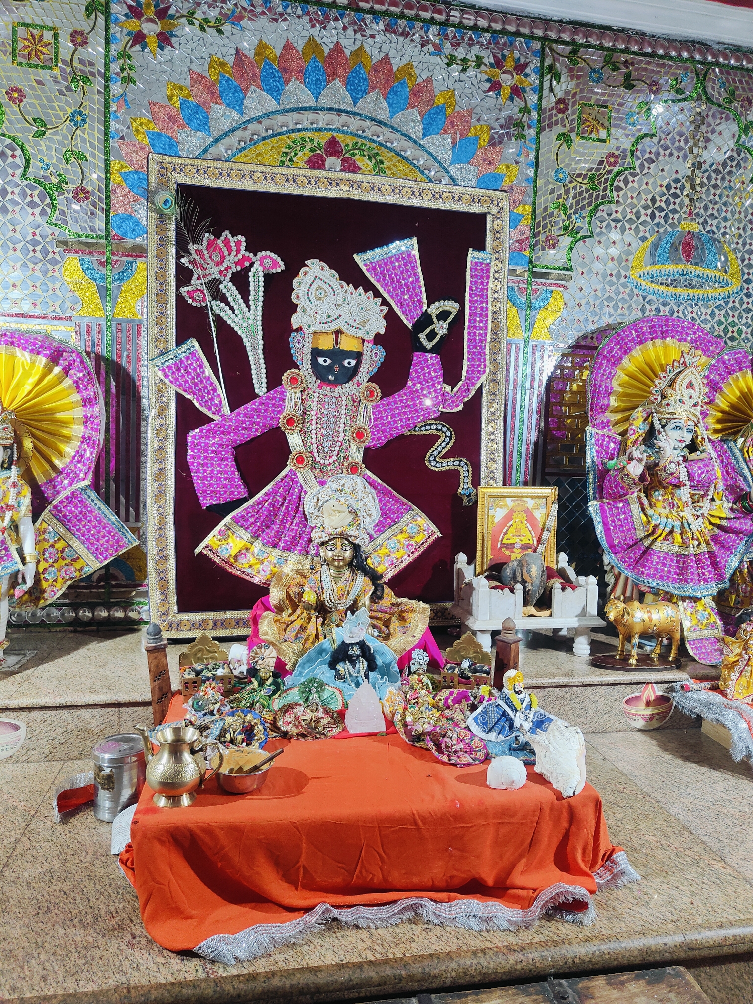 This image depicts an altar in a Hindu temple, likely dedicated to Lord Krishna, located in Gurgaon, India. The central focus is a large, ornate framed picture of a deity, possibly Krishna, adorned with pink and gold clothing and multiple arms. In front of this, a smaller, golden idol of a deity is seated, wearing a white headdress. The altar is covered with an orange cloth and displays various devotional items, including small figurines, a brass jug, a bowl of offerings, and what appears to be a small lamp. Flanking the central deity are two larger, standing idols, also elaborately dressed in pink and gold. The background is decorated with colorful mosaic tiles, featuring floral patterns and shimmering geometric designs. The floor is a speckled granite. The lighting suggests an indoor setting, possibly during evening worship.