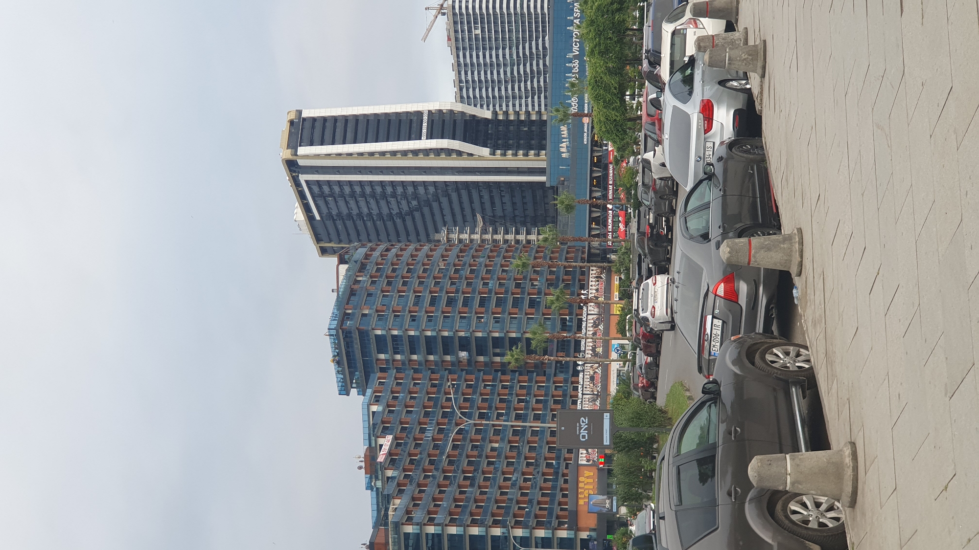 The image depicts a street scene in Batumi, Georgia, featuring a row of parked cars on the right side of the frame, a sidewalk with concrete bollards in the foreground, and several modern high-rise buildings in the background. The buildings are a mix of residential and commercial properties, characterized by their contemporary architecture and large glass facades. Among the visible structures is a distinctive tall, slender building with a white vertical accent. Palm trees are scattered throughout the scene, suggesting a warm climate. Signs and advertisements are visible on the lower levels of some buildings, indicating commercial activity. The sky is overcast, casting a diffused light over the scene. There are no people or active interactions visible in the image, focusing solely on the urban landscape and the parked vehicles.
