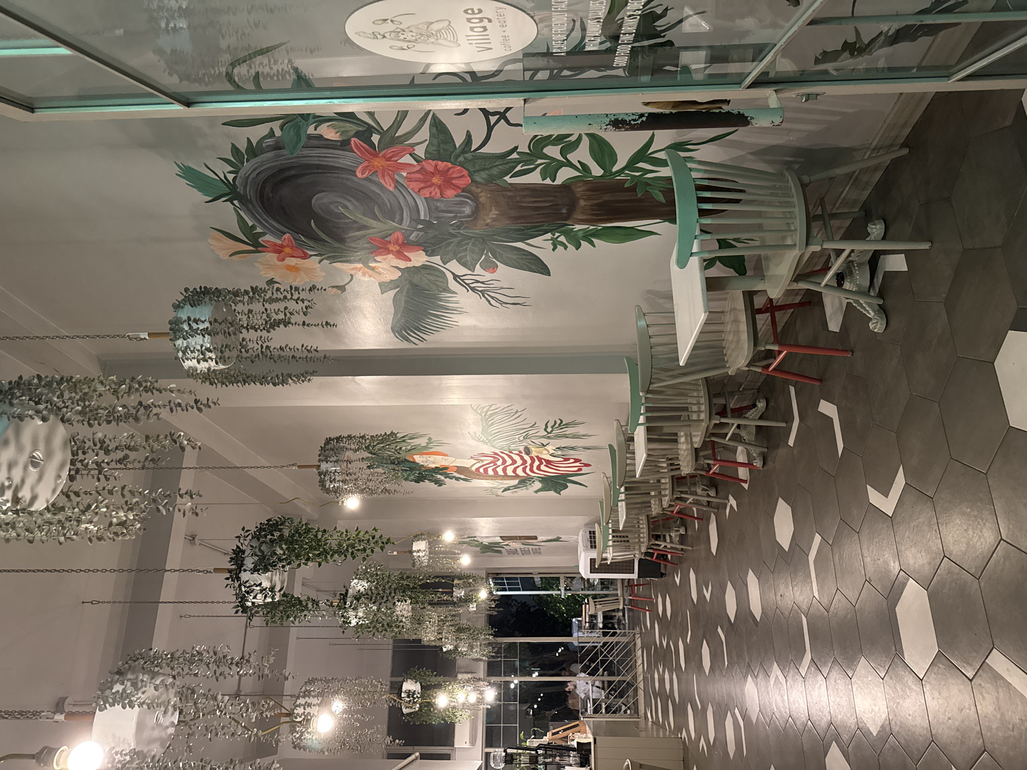 The image depicts the interior of "Village Coffee + Eatery" located in Kadıköy, Turkey. The cafe has a modern, bohemian aesthetic. Hanging plants cascade from the ceiling, creating a lush, green atmosphere. The walls are adorned with a vibrant mural featuring a woman with glasses and red-striped clothing, holding a cup, surrounded by large tropical leaves and flowers.
The flooring is a distinctive hexagonal tile pattern in shades of grey and white. Several tables and chairs are arranged throughout the space. The chairs are predominantly light grey with pastel teal backs, and some have red legs, adding a pop of color. White square tables complement the chairs.
In the background, a counter area is visible with what appears to be a coffee machine and other service equipment. Through a large glass window or door, the exterior of the cafe is partially visible, suggesting a street-level location. A sign on the glass door reads "village coffee + eatery" along with other text, indicating the establishment's name and offerings. A few patrons are seated in the background, suggesting the cafe is open for business. The lighting is warm and ambient, creating an inviting atmosphere.