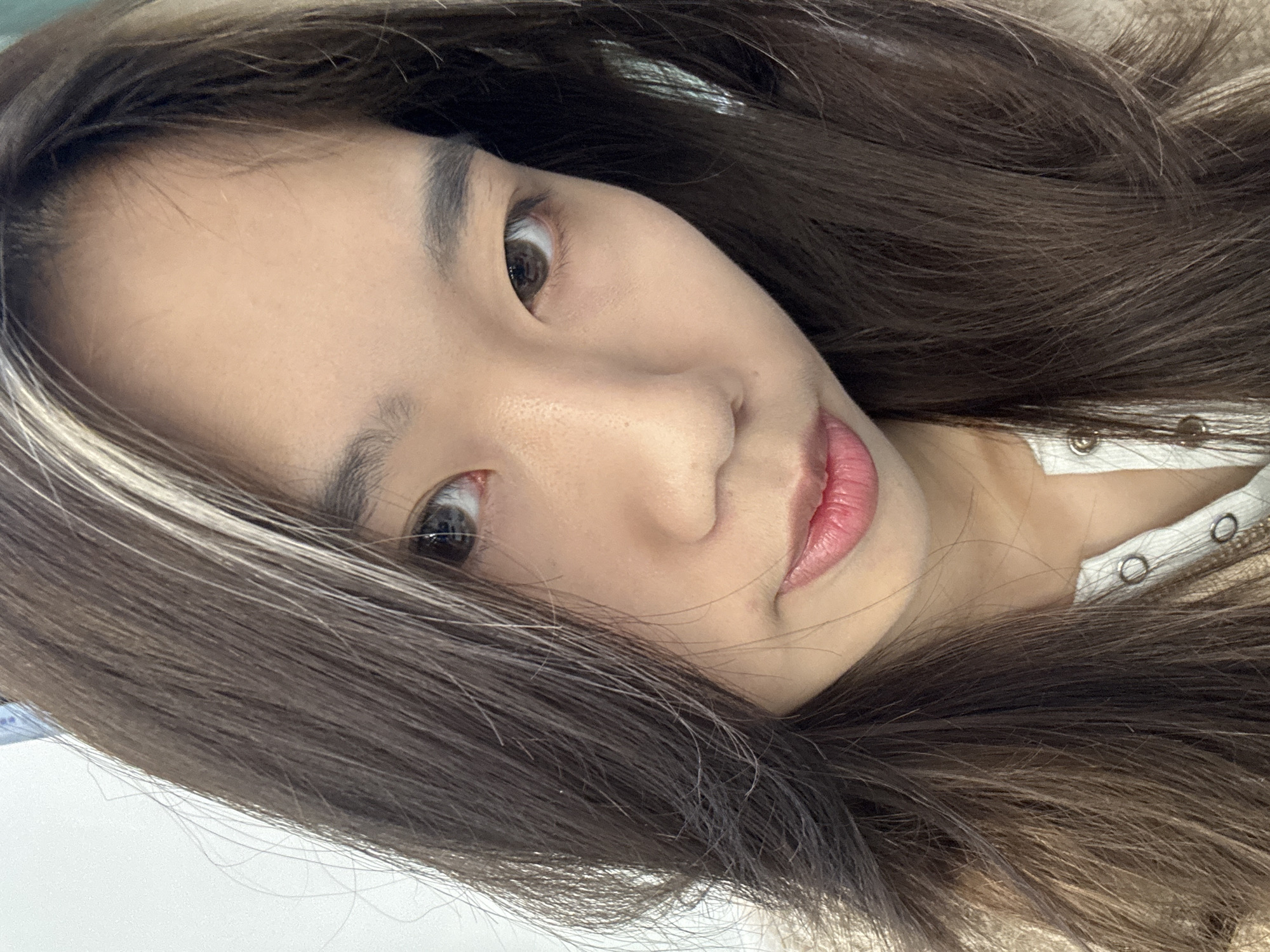 A close-up portrait photograph shows a female individual of East Asian ethnicity looking directly at the camera. Her long, dark brown hair features distinct white/blonde highlights, primarily visible on the viewer's left side. Her eyes are dark brown, with a noticeable redness in the sclera of her right eye (viewer's left). Her eyebrows are dark and well-defined. Her lips are naturally colored in a pinkish-red hue, maintaining a slight, neutral expression. She is dressed in a light-colored top, possibly white or off-white, featuring a placket with several circular metallic buttons. The background is uniformly light and blurred, consistent with an indoor environment with plain walls. The image contains no visual elements to identify a specific location within Nan'an District, China.