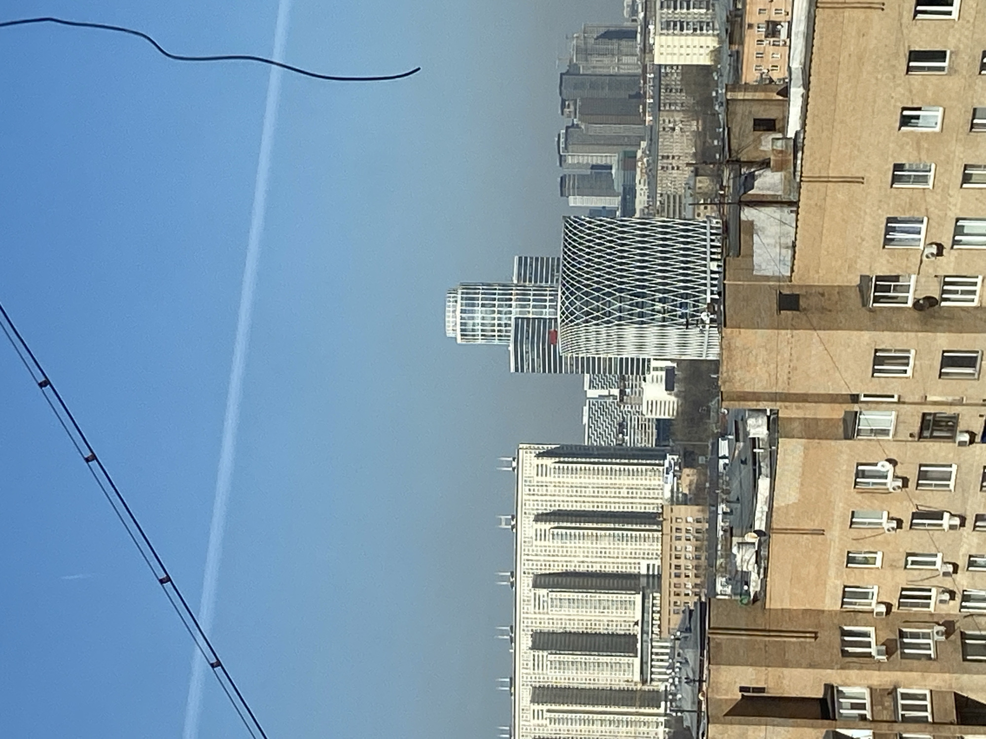 The image shows a partial view of the Moscow cityscape, Russia, under a clear blue sky. Several modern high-rise buildings with glass facades stand next to older, tan-colored apartment buildings with visible rows of windows and air conditioning units. Electrical wires are visible in the foreground against the sky. A jet contrail cuts across the upper left portion of the frame.