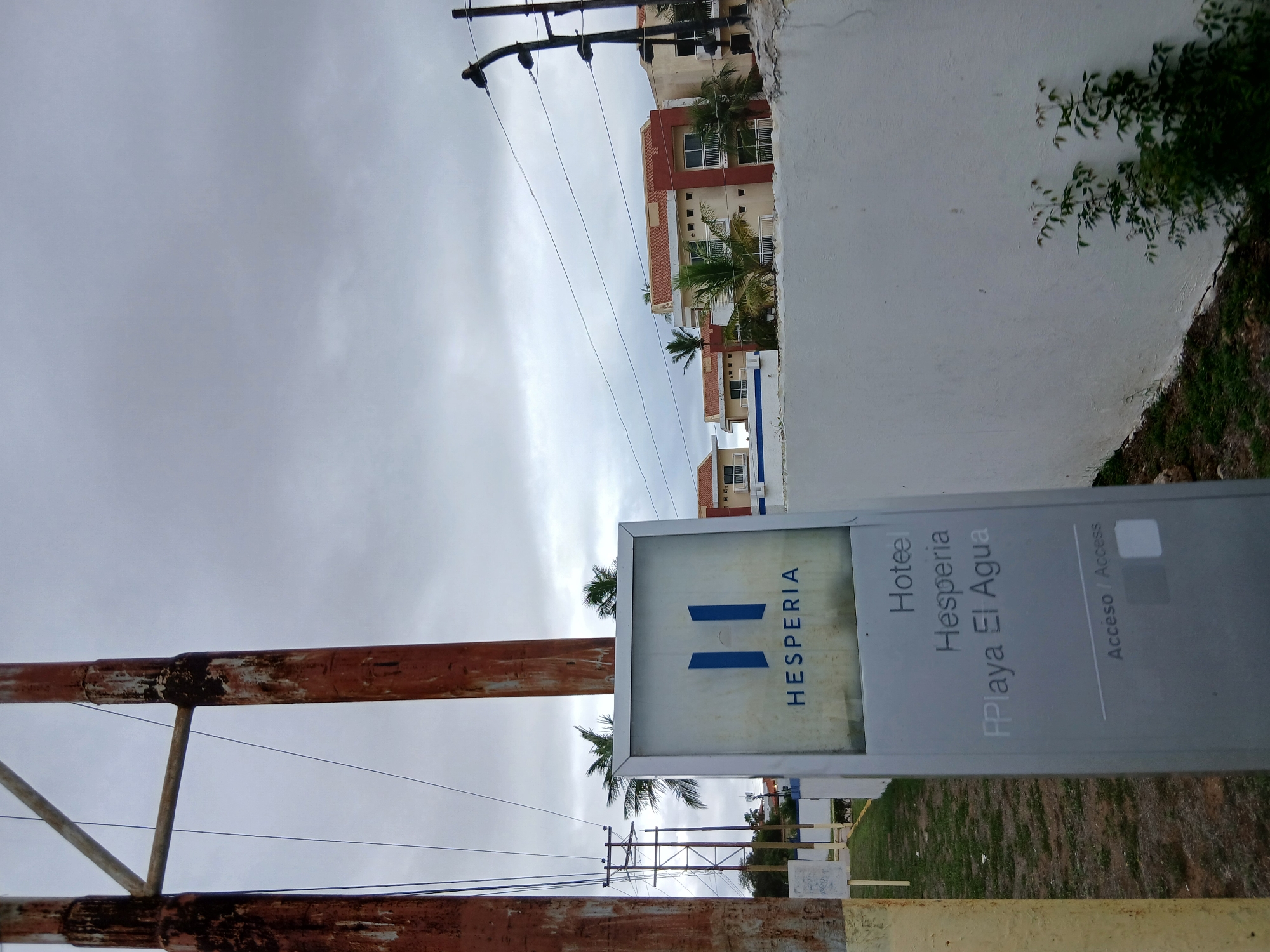 The image shows a sign for "Hotel Hesperia FPlaya El Agua" in Manzanillo, Venezuela. The sign is a light gray rectangular monolith with the Hesperia logo and name at the top. Below this, the text reads "Hoteel Hesperia FPlaya El Agua". At the bottom, it says "Acceso / Access".
The sign is positioned in front of a white wall and to the left of a row of tan and red buildings with orange tile roofs, indicative of a resort or hotel complex. Palm trees are visible in front of the buildings. In the foreground, there are two tall, rusty metal poles with wires running between them and to a utility pole in the background. The sky is overcast with gray clouds. The ground in front of the sign is covered with grass and dirt. The overall scene suggests an outdoor location on a cloudy day.