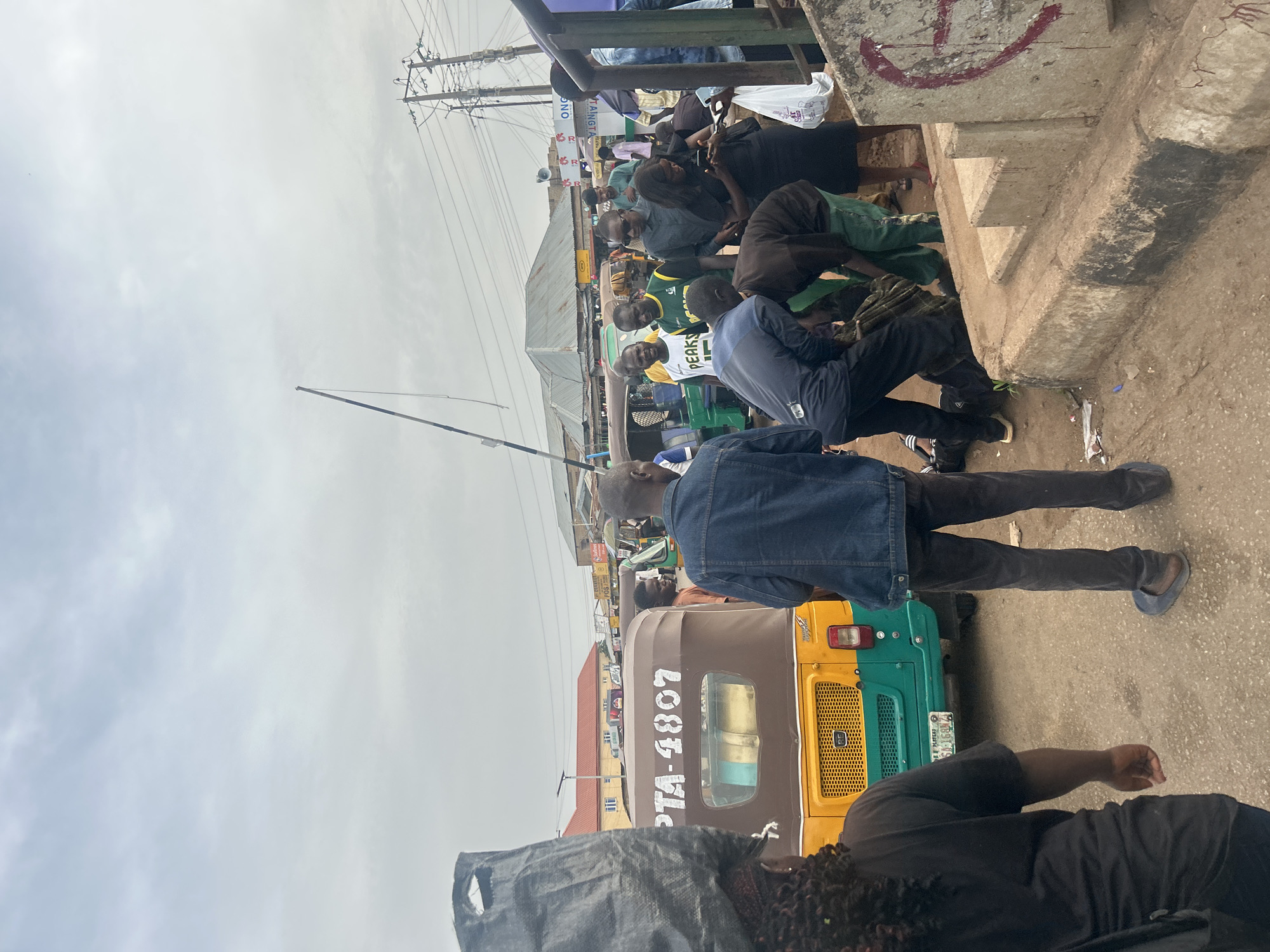 The image shows a street scene in Jos, Nigeria. A crowd of people is gathered near a concrete structure with a red marking. A three-wheeled vehicle, a "Keke Napep" with the number "PTA-4807" on its side, is visible. Utility poles with electrical wires are overhead. Some individuals are dressed in casual clothing. The sky is overcast.