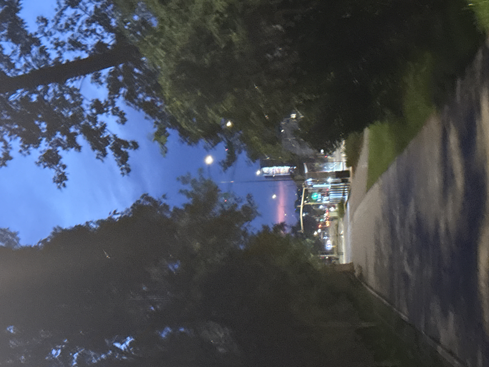 A paved walkway runs along a tree-lined street at dusk. The sky is a deep blue with some clouds, and the lights of the city are visible in the distance. The walkway is bordered by grass and trees on both sides, and there are streetlights and traffic signals visible further down the street. The scene appears to be a quiet urban area at the end of the day.