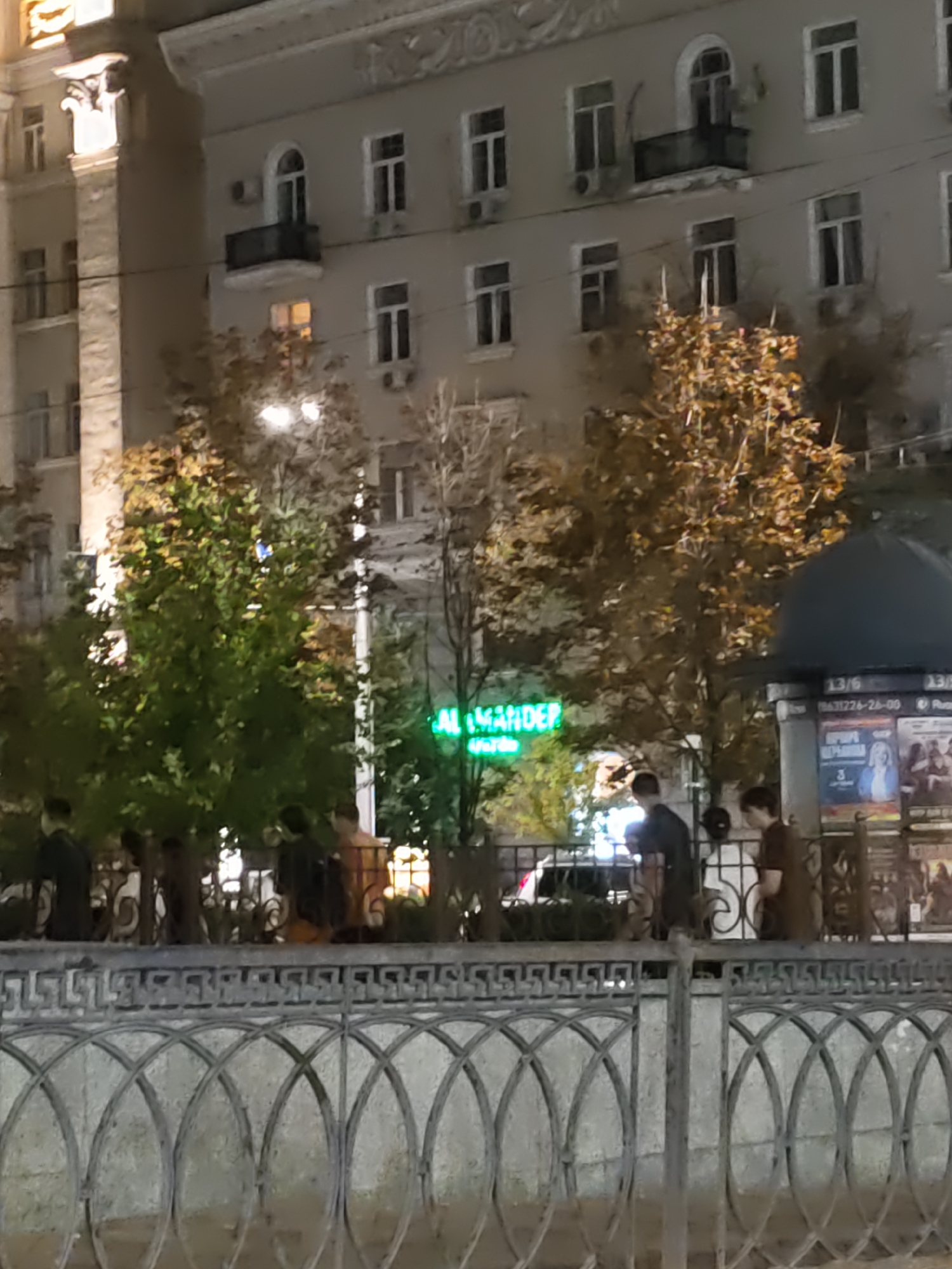 The image shows a nighttime street scene in Rostov-on-Don, Russia, likely on Bolshaya Sadovaya Street. In the background, a multi-story building with neoclassical architectural elements is visible, illuminated by streetlights and interior lights from its windows. Trees with autumn foliage are positioned in front of the building, with some illuminated by streetlights. A green neon sign with the word "ALEXANDER" is prominently displayed on one of the trees. In the foreground, a decorative metal fence runs horizontally across the image. Several individuals are standing behind the fence, some appearing to be in conversation. A kiosk or information stand with posters is visible on the right side of the image. The overall atmosphere is urban and nocturnal.