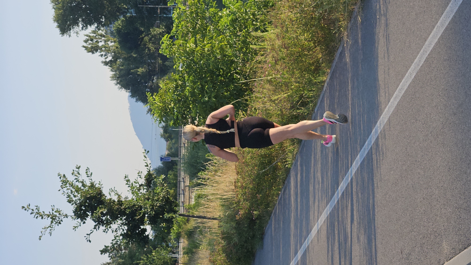 A blonde woman with a braid is running on a paved path with a white line marking the lane. She is wearing a black sports bra and black shorts. The path is bordered by dry grass and green bushes. In the background, there are trees, a hill, and a clear blue sky. The scene appears to be outdoors, possibly in a park or a designated running track, during daylight. There is no specific visual information to confirm the location as Prešov, Slovakia.