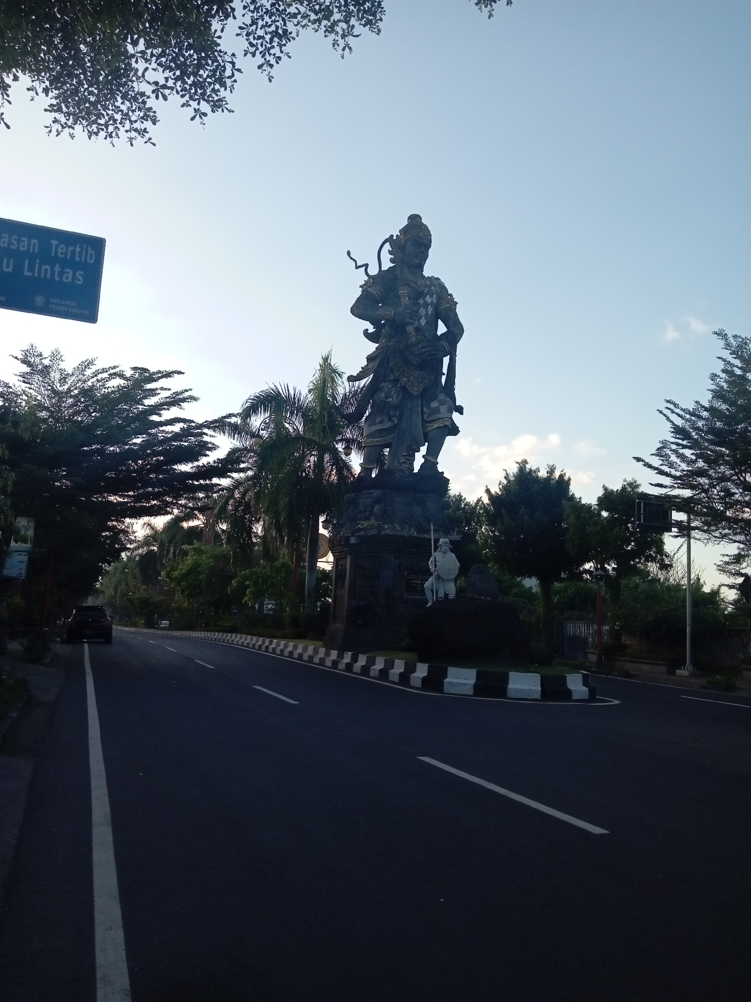 An outdoor, low-angle photograph captures a roadway leading to a central roundabout or median featuring a prominent statue, taken during what appears to be early morning or late afternoon.
In the center of the image, a towering, dark-colored statue of a multi-armed warrior figure stands atop a stone pedestal. The figure is adorned with intricate details and headwear, holding various implements including a bow and a serpent-like object. At the base of this large statue, a smaller, lighter-colored statue of a male warrior, possibly holding a spear and a round shield, is visible.
To the left of the scene, a two-lane asphalt road recedes into the distance, with a solid white line marking the shoulder and a dashed white line separating the lanes. A dark-colored car is partially visible parked or driving on the left side of the road further back. A blue rectangular road sign is positioned on the left, partially obscured by tree leaves, displaying white text that includes "Tertib", "Lintas", and "SATLANTAS POLRES GIANYAR".
The road on the right is bordered by a raised median or curb constructed with alternating black and white checkered blocks. Various trees, including tall palm trees and dense broadleaf foliage, line both sides of the road and form a backdrop for the statue. Some tree leaves are silhouetted against the sky in the top left corner.
The sky is a clear, light blue with scattered white clouds, suggesting an ambient light condition. The foreground road and lower parts of the scene are cast in soft shadow, contrasting with the brighter sky. No people are discernible in the image. The location is Gianyar, Indonesia, based on the road sign.