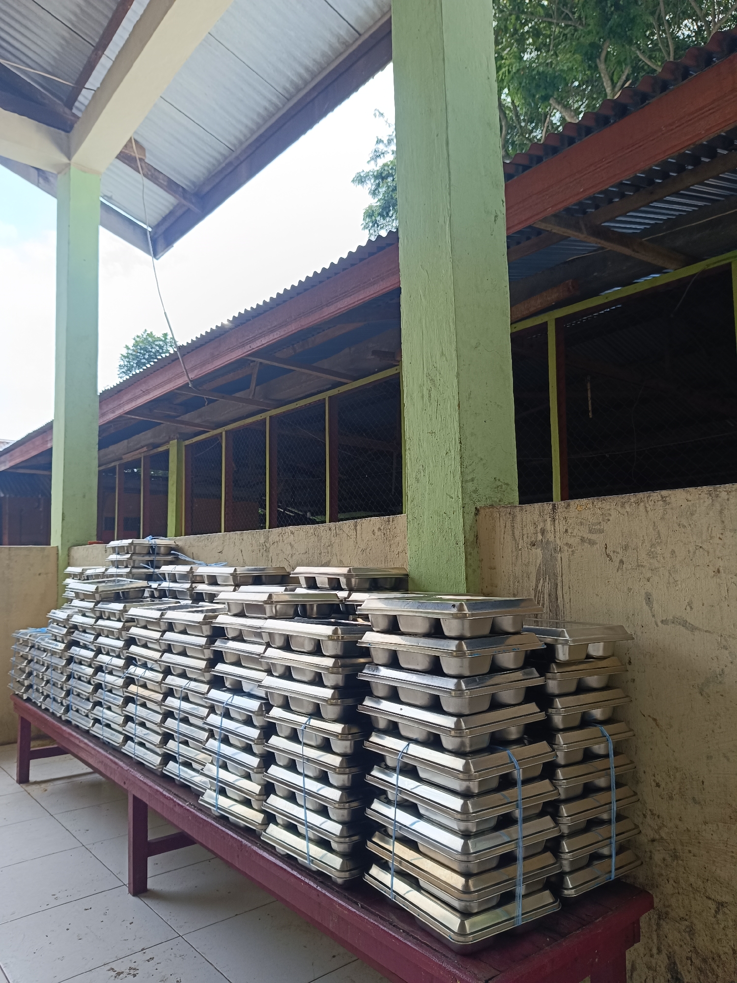 A large stack of aluminum trays sits on a maroon bench outside a building in Lhokseumawe, Indonesia. The building has a corrugated iron roof and light green columns. A section of the building is enclosed with wire mesh. The trays are stacked neatly and tied with blue plastic straps. The floor is tiled and the wall is a light yellow color. The sky is visible in the background.