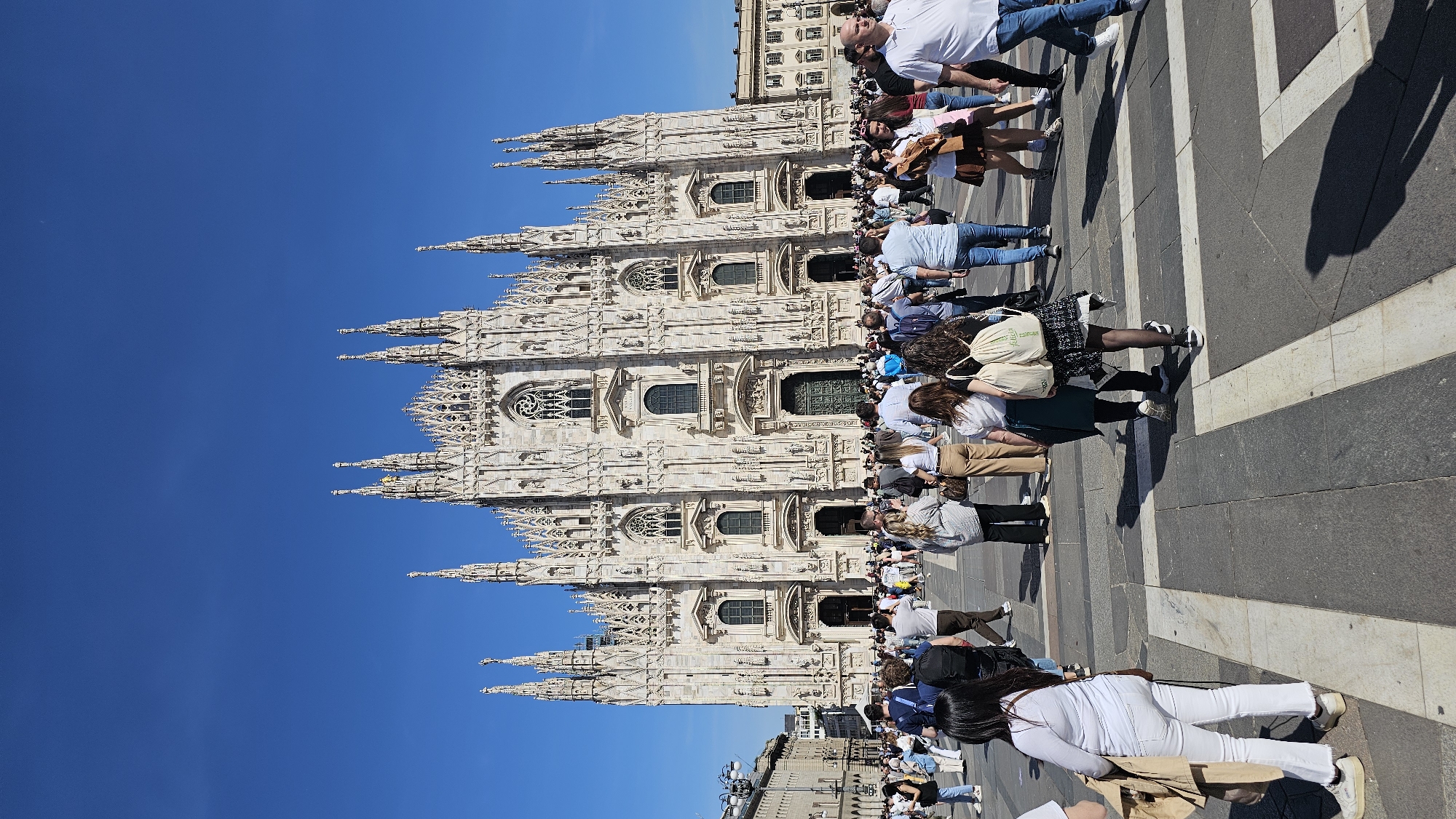 The image depicts the Milan Cathedral (Duomo di Milano) in Milan, Italy, from a ground-level perspective. The cathedral's facade is visible, showcasing its intricate Gothic architecture. Numerous people are present in the foreground, seemingly tourists and visitors. The sky is clear and blue. The ground exhibits a pattern of gray and white stones, forming a distinct design, possibly part of the piazza. People are walking and standing, and some are interacting with each other.