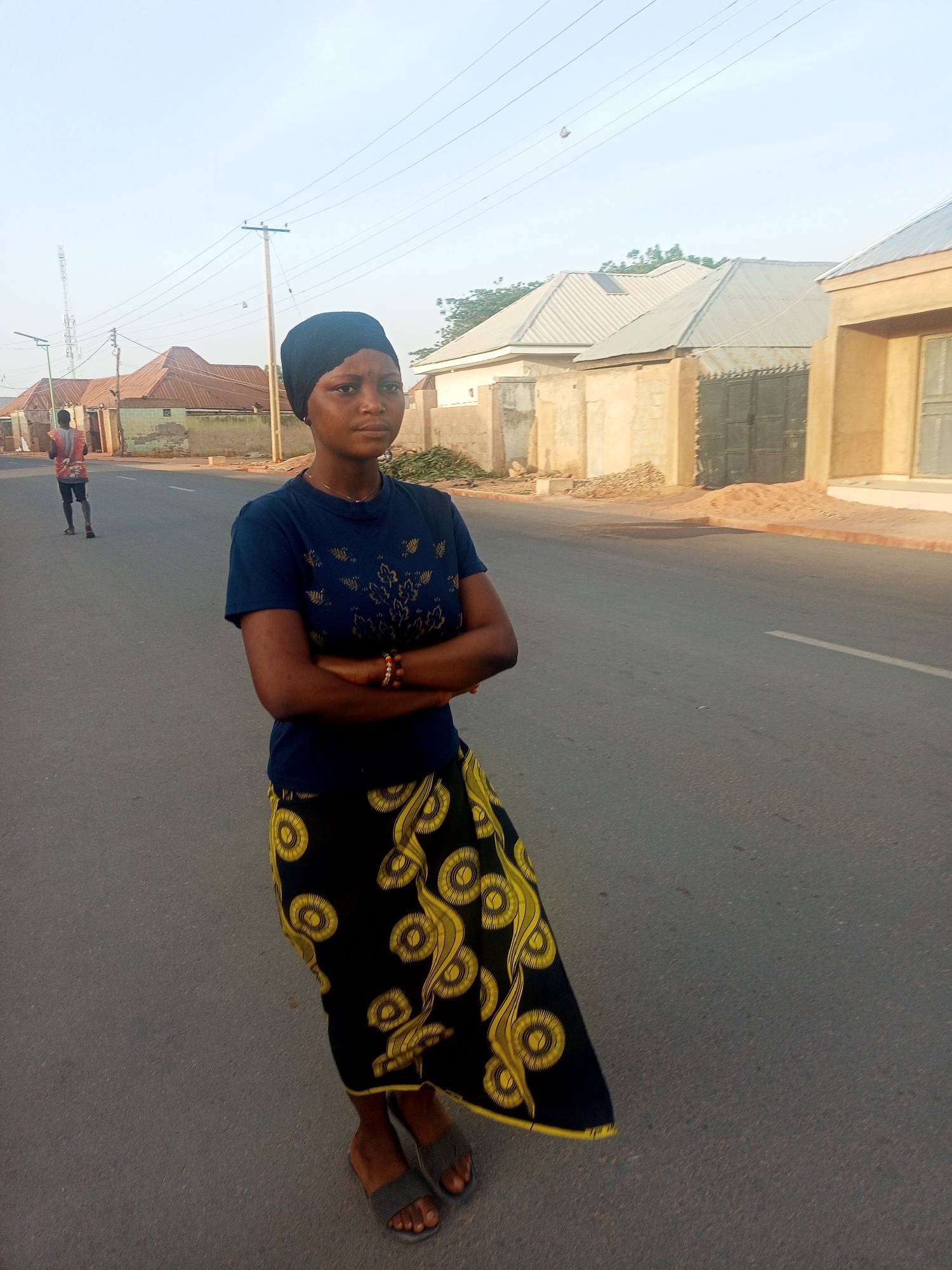 A young woman stands in the middle of a paved road in Tempure, Nigeria. She is facing forward with her arms crossed. She wears a dark blue t-shirt with gold-colored designs, a long, patterned black and gold skirt, and dark sandals. Her head is covered with a dark scarf. A bracelet is visible on her right wrist. In the background, there are residential buildings on either side of the road. A man is walking away in the distance on the left side of the road. Overhead power lines are visible against a light sky. The lighting suggests it is daytime.