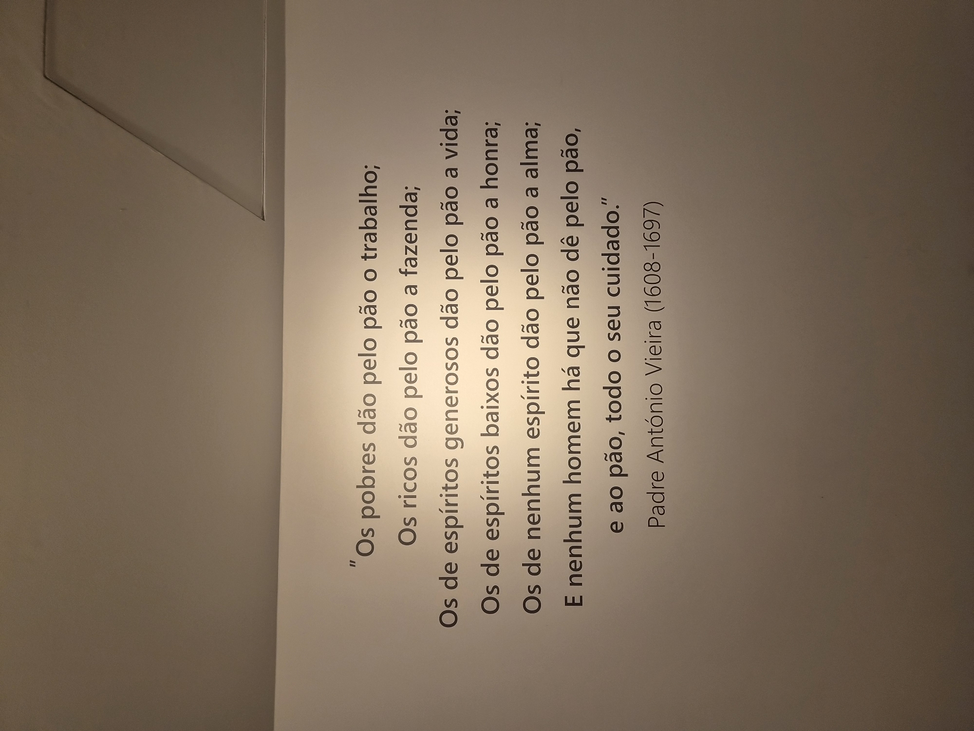 The image is a vertical, close-up shot of a wall with text printed on it. The text is in Portuguese and appears to be a quote. The quote is attributed to "Padre António Vieira (1608-1697)". The wall is a neutral, light color, with a subtle shadow or architectural detail visible in the upper left portion of the frame. The lighting is even, suggesting an indoor setting. The text itself is the primary subject. The quote, when translated, discusses how different types of people provide for "bread" (representing sustenance or livelihood), with the poor contributing labor, the rich their property, the generous their lives, the mean-spirited their honor, and the spiritless their souls. The final line states that no man exists who doesn't give his care to bread. The text is printed in a dark, sans-serif font. The image is likely taken in Seia, Portugal, as indicated by the location context, though no specific visual cues in the image directly point to this location.