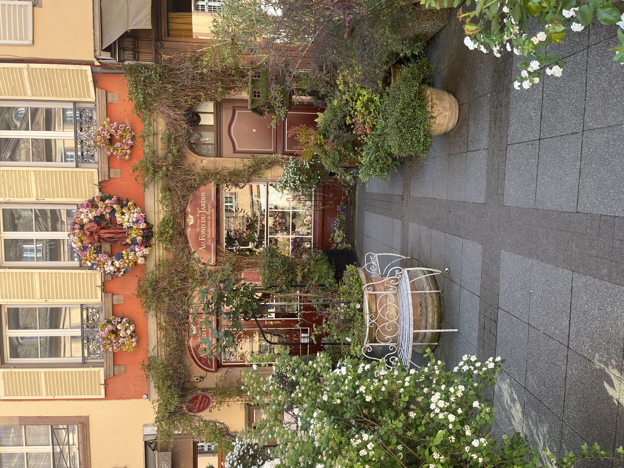 The image captures the entrance to "Au Fond Du Jardin," a tea room and brunch spot in Strasbourg, France. The building is adorned with floral wreaths, climbing vines, and potted plants. A white wrought-iron bench sits near the entrance, surrounded by blooming bushes. The walkway leading to the doorway is paved with gray tiles. The sign indicates the business has been established since 1999. The architecture is traditional Alsatian, with orange-painted walls and shuttered windows.