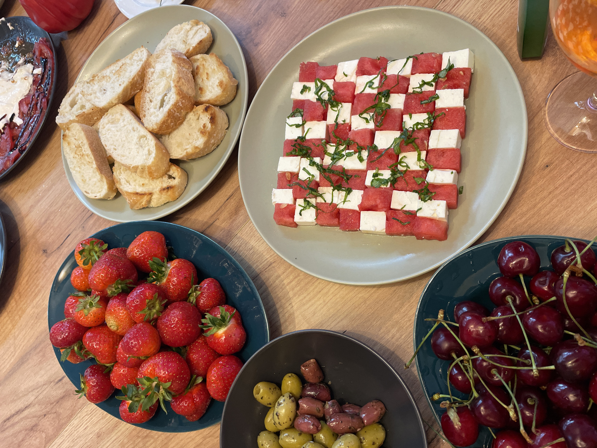 On a wooden table, a variety of food items are arranged on plates. In the foreground, a dark blue plate is filled with ripe cherries. To the left, a dark grey bowl contains green and purple olives. Above the olives, a dark blue plate holds a pile of fresh strawberries. To the right of the strawberries, a light grey plate features slices of toasted bread. Further to the right, a light grey plate showcases a checkerboard pattern of cubed watermelon and white cheese, garnished with chopped green herbs. In the background, another plate holds a dish with sliced tomatoes, cheese, and a drizzle of dark sauce. The scene appears to be set indoors in an unknown location.