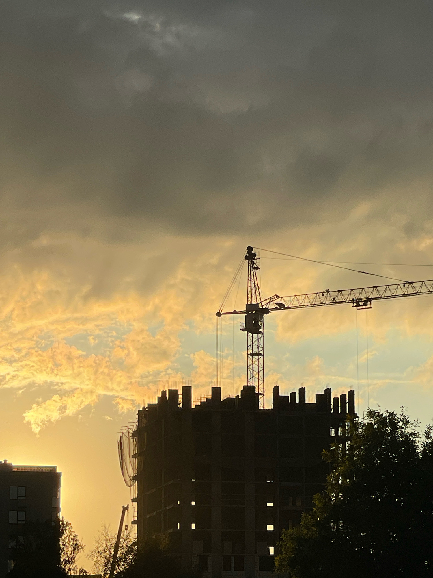 The image depicts a construction site in Izhevsk, Russia, at dusk. A large construction crane is silhouetted against a dramatic sky filled with heavy, dark clouds interspersed with areas of bright, yellowish light, suggesting the sun is low on the horizon. In the foreground, the dark silhouette of a partially constructed building dominates the center of the frame. Its unfinished upper floors show exposed concrete rebar and empty window openings. To the left, another building, appearing more complete with visible windows, is also silhouetted against the bright sky. Trees with dark foliage are visible in the lower right portion of the image, further silhouetted by the fading light. The overall scene conveys a sense of ongoing development and the atmospheric conditions of an evening sky.