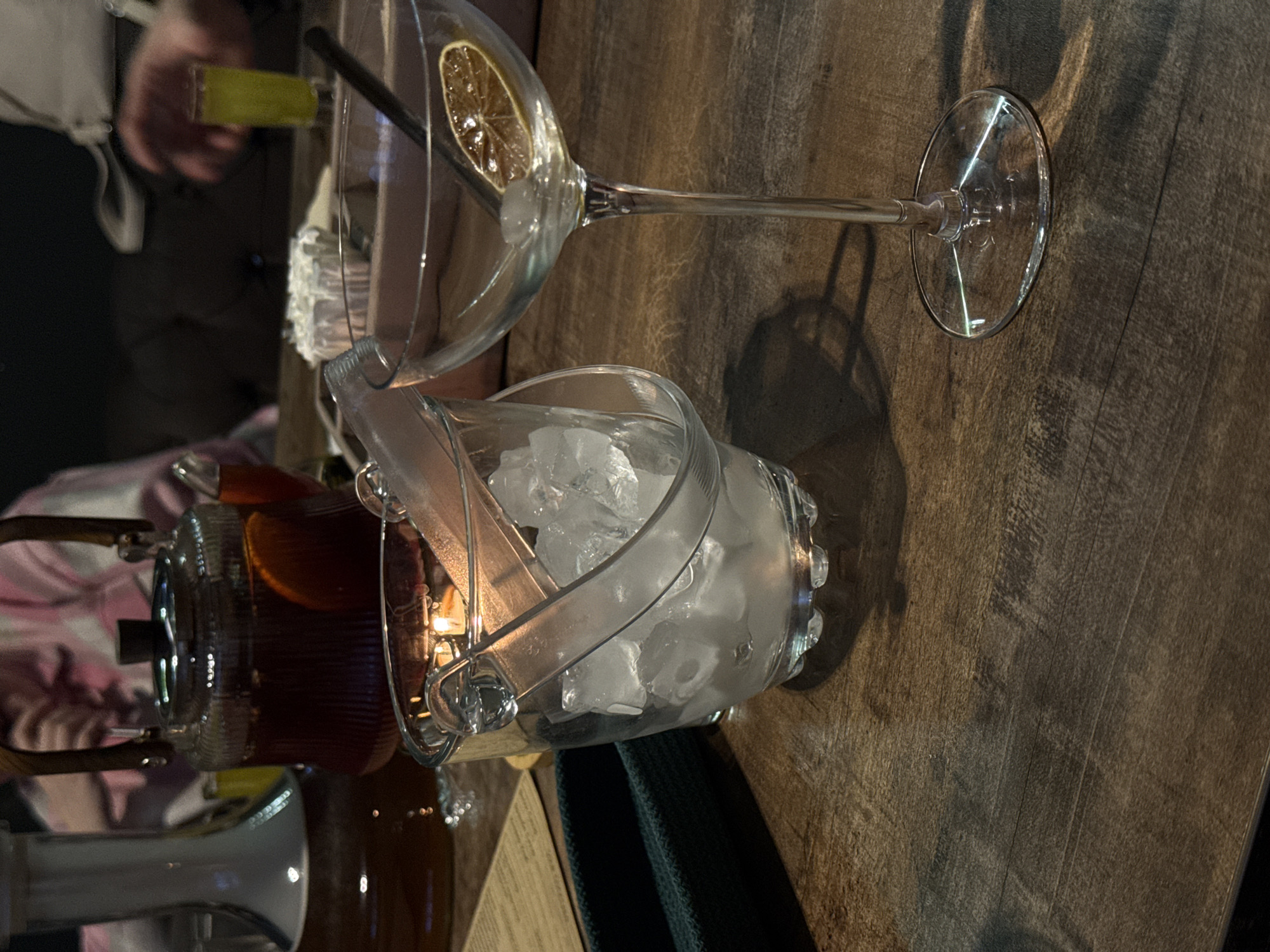 The image captures a table setting, likely in a bar or restaurant. The table has a wooden surface. There is a tall glass with a sliced lemon/orange garnish, a black straw, and possibly ice. An ice bucket filled with ice cubes is positioned near the glass. A dispenser containing a dark-colored liquid, possibly iced tea or a mixed drink, is present. The overall scene suggests a cocktail or beverage preparation setup. The location in Chaikovsky, Russia is not discernible from the image itself. There are two people visible in the image. One person is wearing a pink and white checkered shirt, and the other is wearing a gray and black shirt. Both people are reaching towards the table.