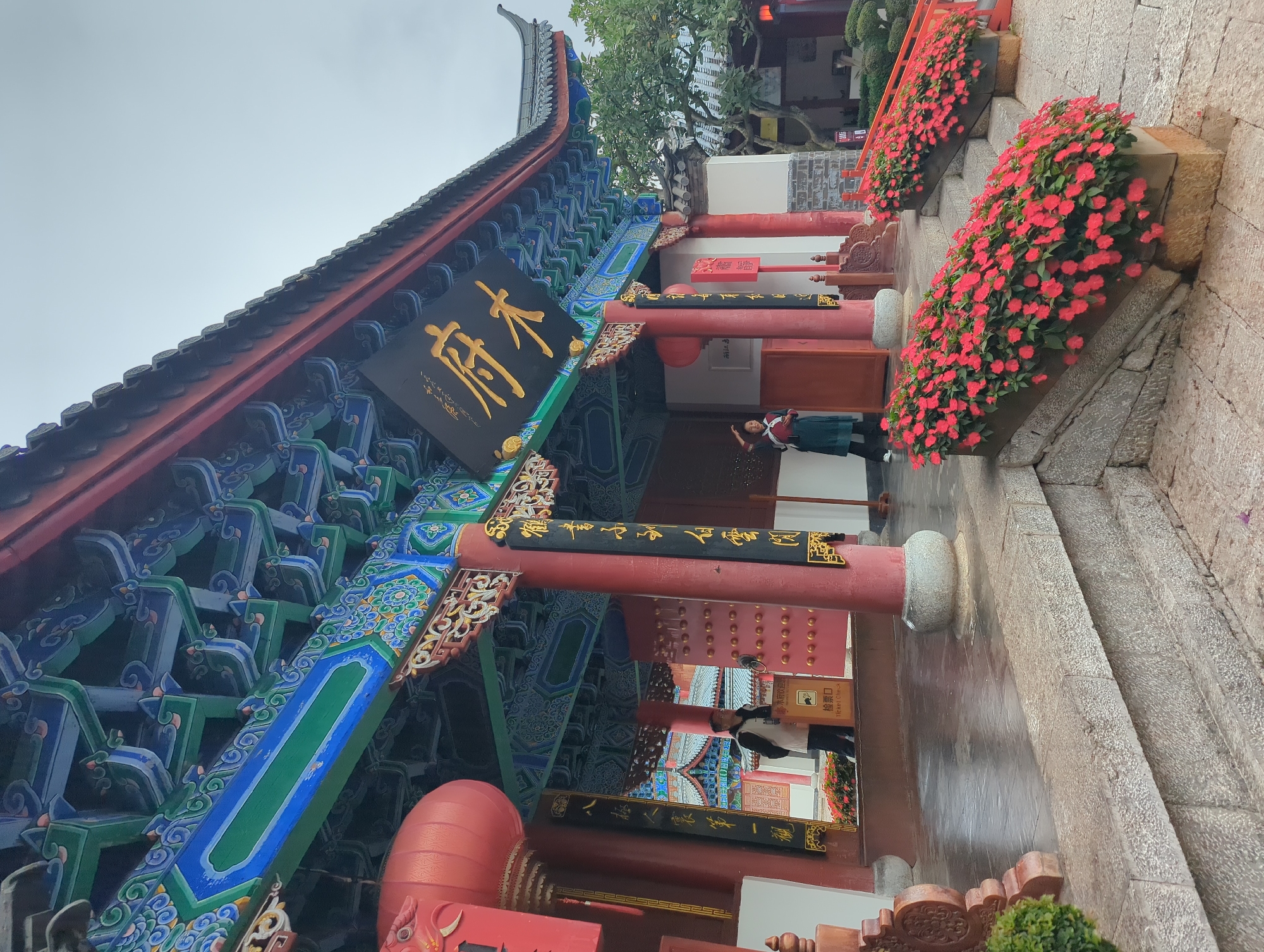 The image captures the entrance to a traditional Chinese building in Gucheng District, China. The building features a brightly painted roof with intricate blue, green, and red decorative patterns. A large black sign with golden Chinese characters is prominently displayed above the entrance. Red lanterns hang on either side of the entrance, and red pillars support the eaves of the roof.
Two individuals are visible at the entrance. On the left, a person in a light-colored jacket stands near a ticket check sign. On the right, a person in a dark green outfit is posing with their hand on their head, looking towards the left.
The foreground is adorned with planters filled with vibrant red flowers, cascading down the stone steps leading to the building. The ground is paved with stone, and parts of it appear wet, reflecting the overcast sky. In the background, more traditional Chinese architecture is visible, along with lush green trees and foliage. The overall scene conveys a sense of cultural heritage and natural beauty.