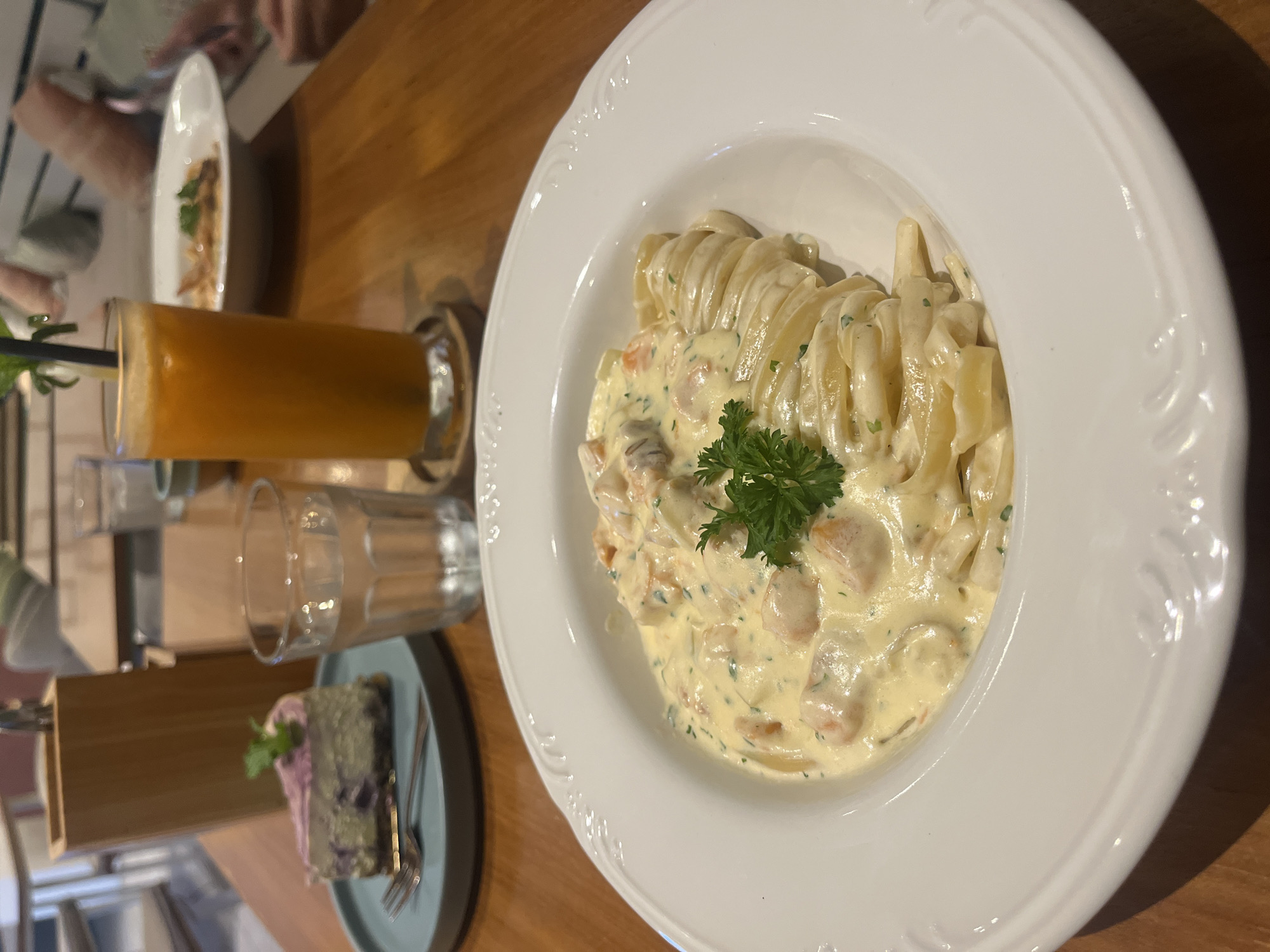 The image presents a vibrant dining scene, likely inside a restaurant or cafe, with food and beverages arranged on a warm wooden table. The central focus is a large, elegant white ceramic bowl containing a generous serving of pasta. The pasta, resembling tagliatelle or fettuccine, is coated in a rich, creamy, light yellow sauce, interspersed with visible pieces of what appears to be shrimp or salmon, and finely chopped green herbs. A fresh sprig of parsley garnishes the top of the dish. The white bowl features an intricately embossed decorative rim.
To the left of the pasta, a tall glass holds an orange-colored juice, complete with a black straw and a small green garnish, possibly a mint leaf or a slice of citrus. Beside the juice, an empty clear drinking glass is placed on the table. Further into the foreground and to the left, a slice of cake rests on a small, light blue/grey ceramic plate. The cake itself exhibits a distinct purple or greyish color, topped with a streak of pinkish-purple frosting and a tiny green leaf for garnish. A silver fork is positioned next to the cake.
In the blurred background, indistinct hands and arms of other individuals suggest additional diners at the table. The setting features warm lighting, and the wooden table surface contributes to a cozy ambiance. The scene is located in Kuta Selatan, Indonesia. There is no visible text present in the image.