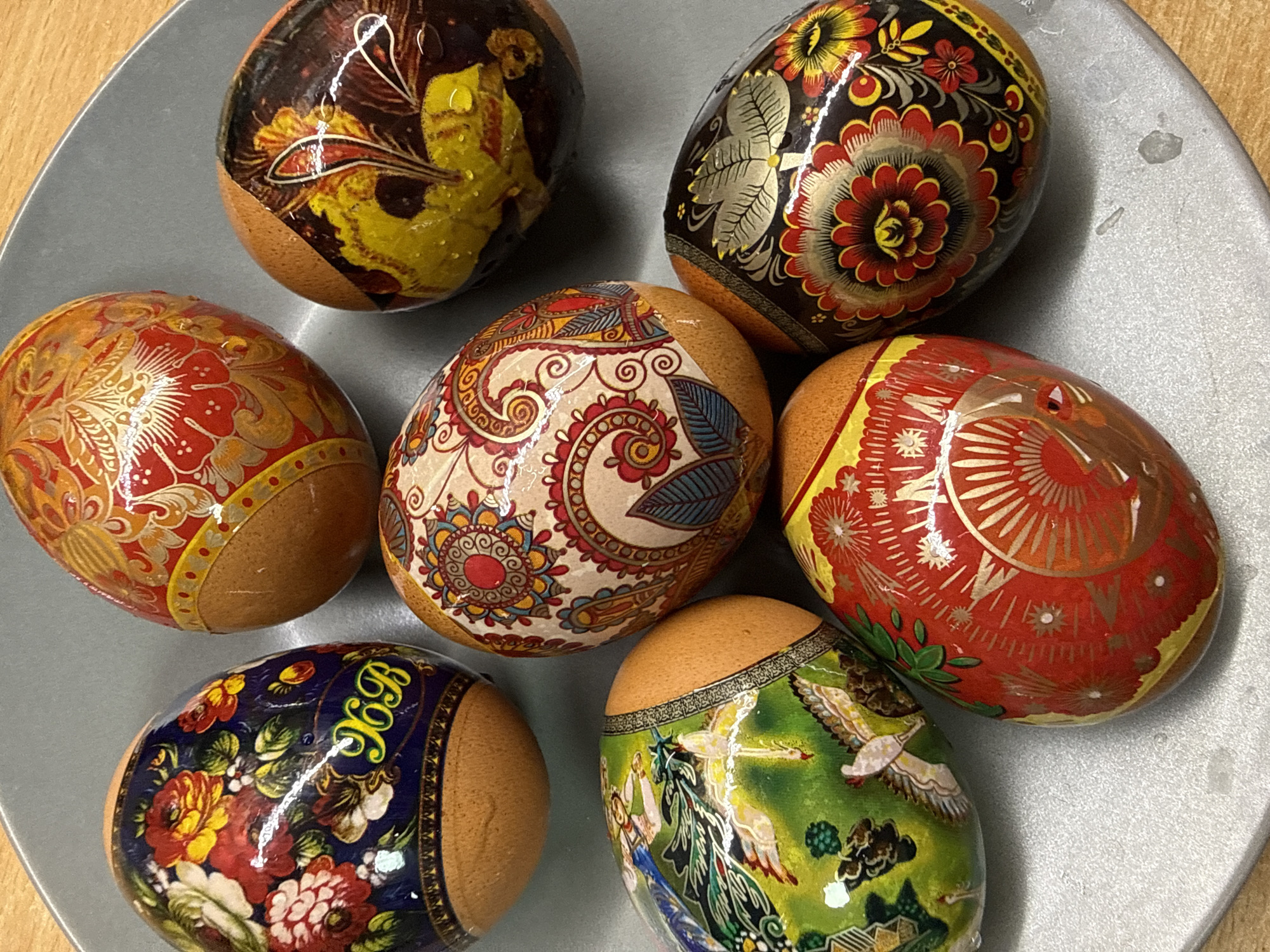 A top-down shot shows seven decorated eggs on a gray plate. The eggs feature a variety of intricate designs, including floral patterns, paisleys, and depictions of people and animals. The base of each egg appears to be a natural brown color. The plate has minor surface imperfections. The location and city are unknown.