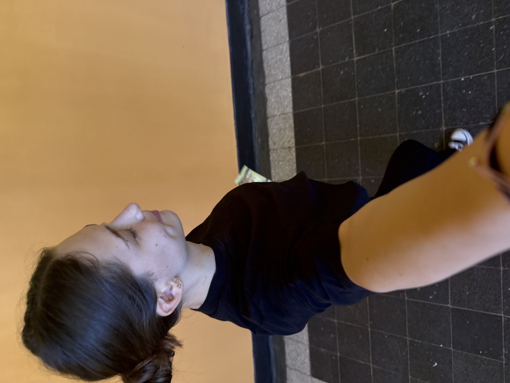 A young woman is taking a selfie in what appears to be a hallway. She is wearing a black long-sleeved shirt and has her dark hair pulled back in a bun. She is wearing small stud earrings. The background consists of a textured orange wall above a dark baseboard, and below that, a floor tiled with dark grey and black squares. The image does not provide any specific information about a location in the city of Sofia, Bulgaria.