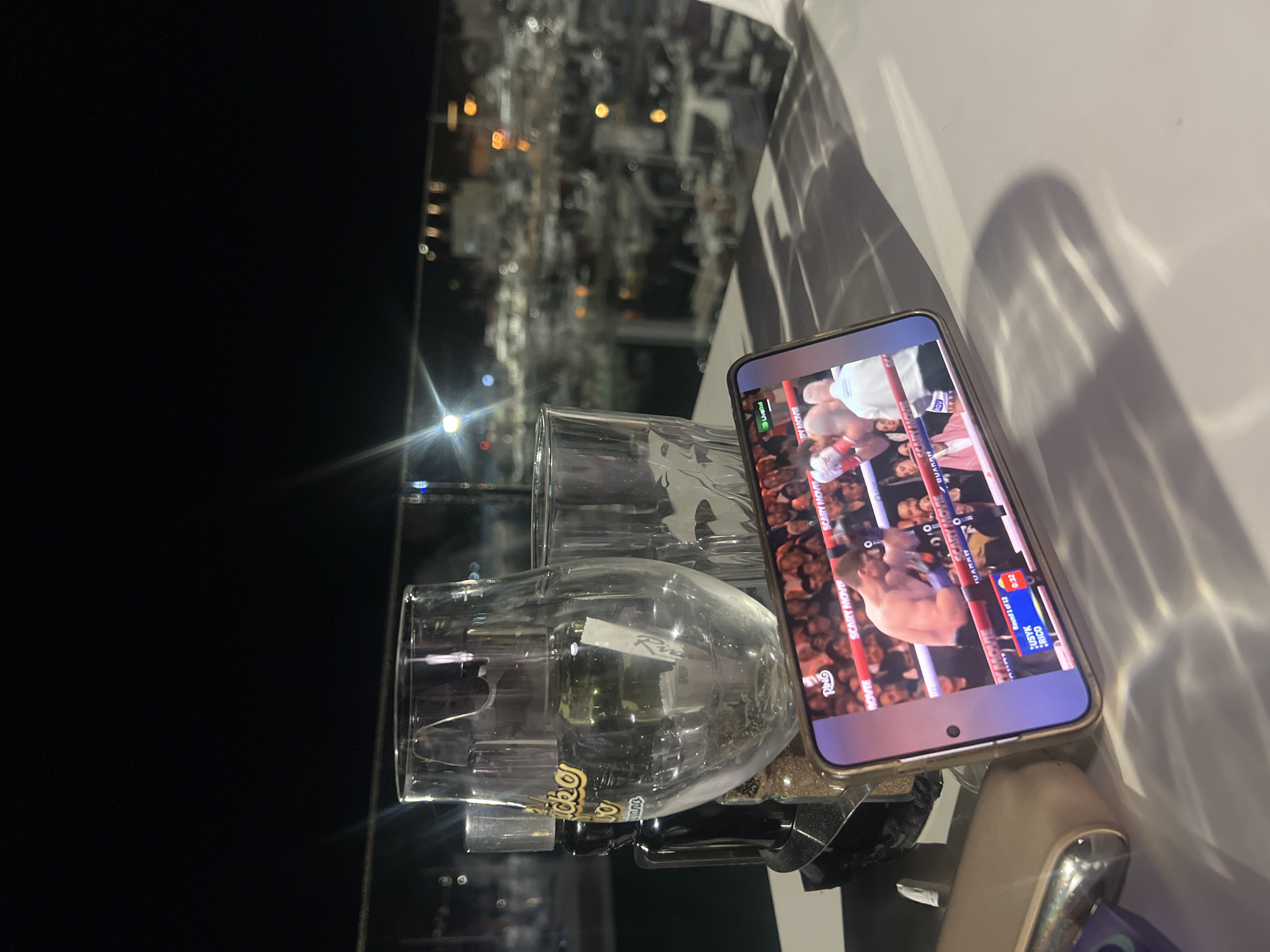 The image shows a low-angle, indoor scene at night. Two empty, clear glass beer steins are positioned near the center of the frame. A smartphone is placed on the right side, displaying a boxing match. The screen shows two boxers in action. A crowded audience is visible in the background of the phone screen. The room reflects a blurred cityscape through a glass panel behind the glasses, with various lights indicating a nighttime urban environment. Light reflecting off the steins creates glare. The image was likely taken in Budva, Montenegro.