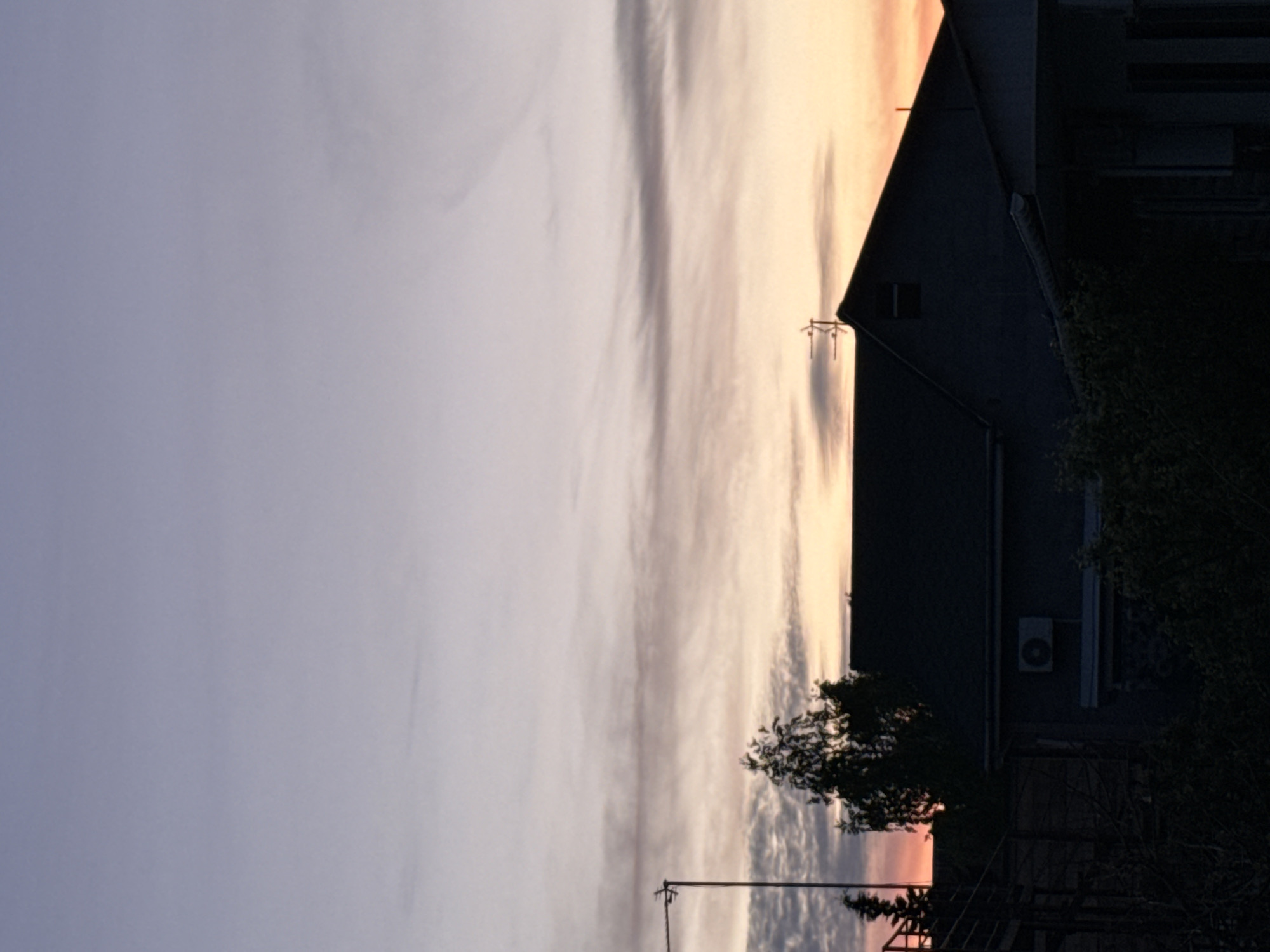 The image depicts a dusk or dawn sky in Береговой сельский совет, Ukraine, with pastel colors transitioning from pale orange to muted purple. Wispy clouds stretch across the sky. In the lower right, a darkened building is visible, featuring a sharply angled roof. An air conditioning unit is mounted on the building's exterior. A tree with sparse foliage sits in front of the building, obscuring some of the structure. An electrical pole is situated near the bottom center of the image. The scene appears still and quiet, suggesting a residential or suburban location.