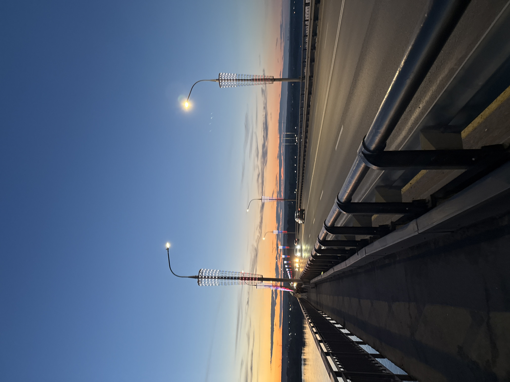 This is an evening scene on a bridge in Perm, Russia, featuring distinctive street lamps. The bridge is paved and has metal railings on the right side. A row of street lamps with cylindrical, illuminated structures along the bridge extends into the distance. The sky is a gradient of blue to orange, suggesting either sunrise or sunset. There are cars visible on the bridge. The image is taken at eye level. The water is visible under the bridge, and cityscape is visible at the far end of the bridge.