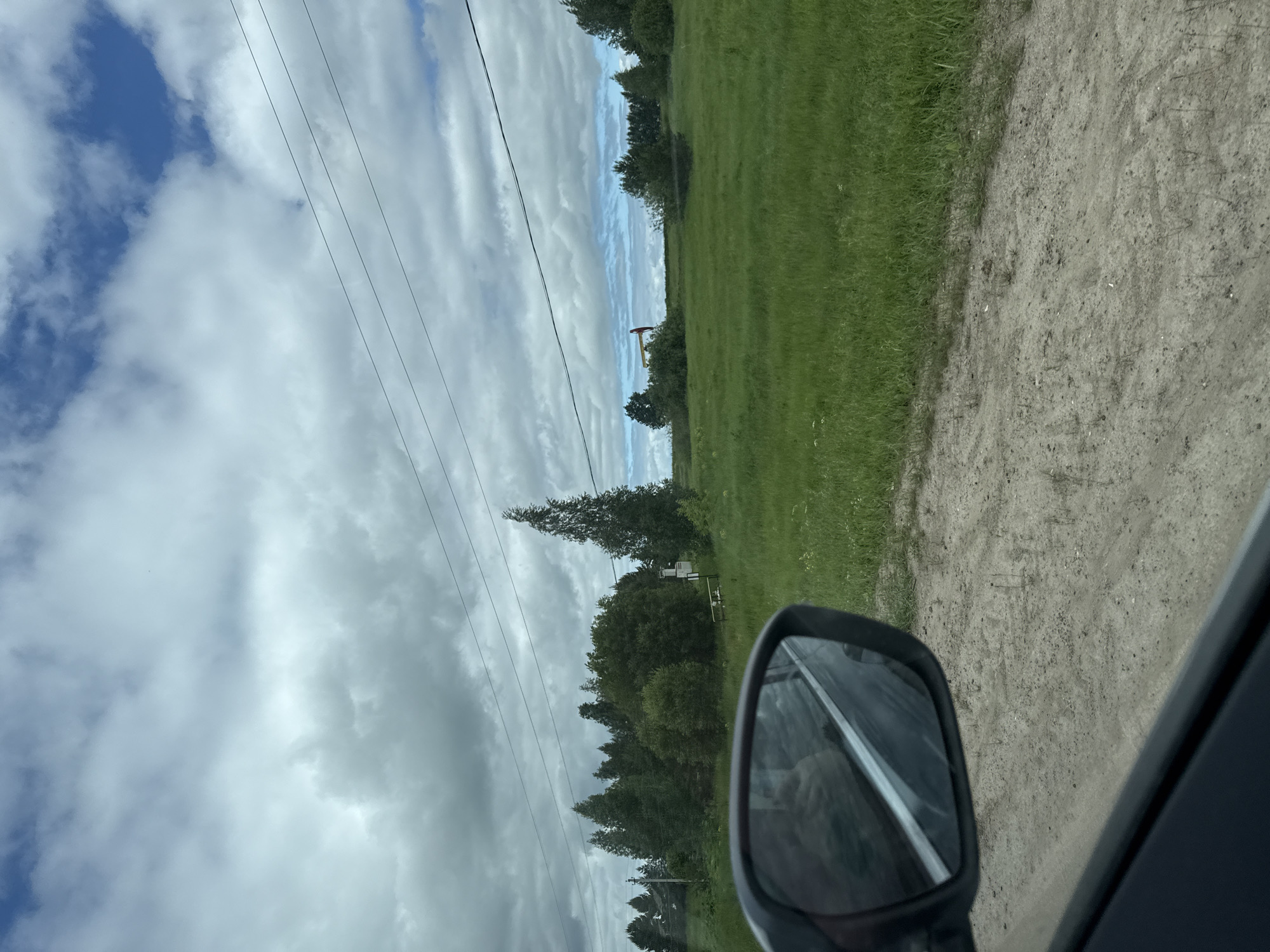 The image shows a view from a car, looking out the window and into the side mirror, with a landscape of green fields and trees under a cloudy sky. In the far distance, there is a yellow structure with a red element, possibly industrial equipment or machinery. The side mirror reflects a person's face and the interior of the car. The ground in the foreground appears to be a dirt or gravel road. The sky is partly cloudy with patches of blue visible. The scene is likely located in a rural area, as suggested by the open fields and natural vegetation. There is no specific information to indicate the location within the city of Krasnoyarsk, Russia.