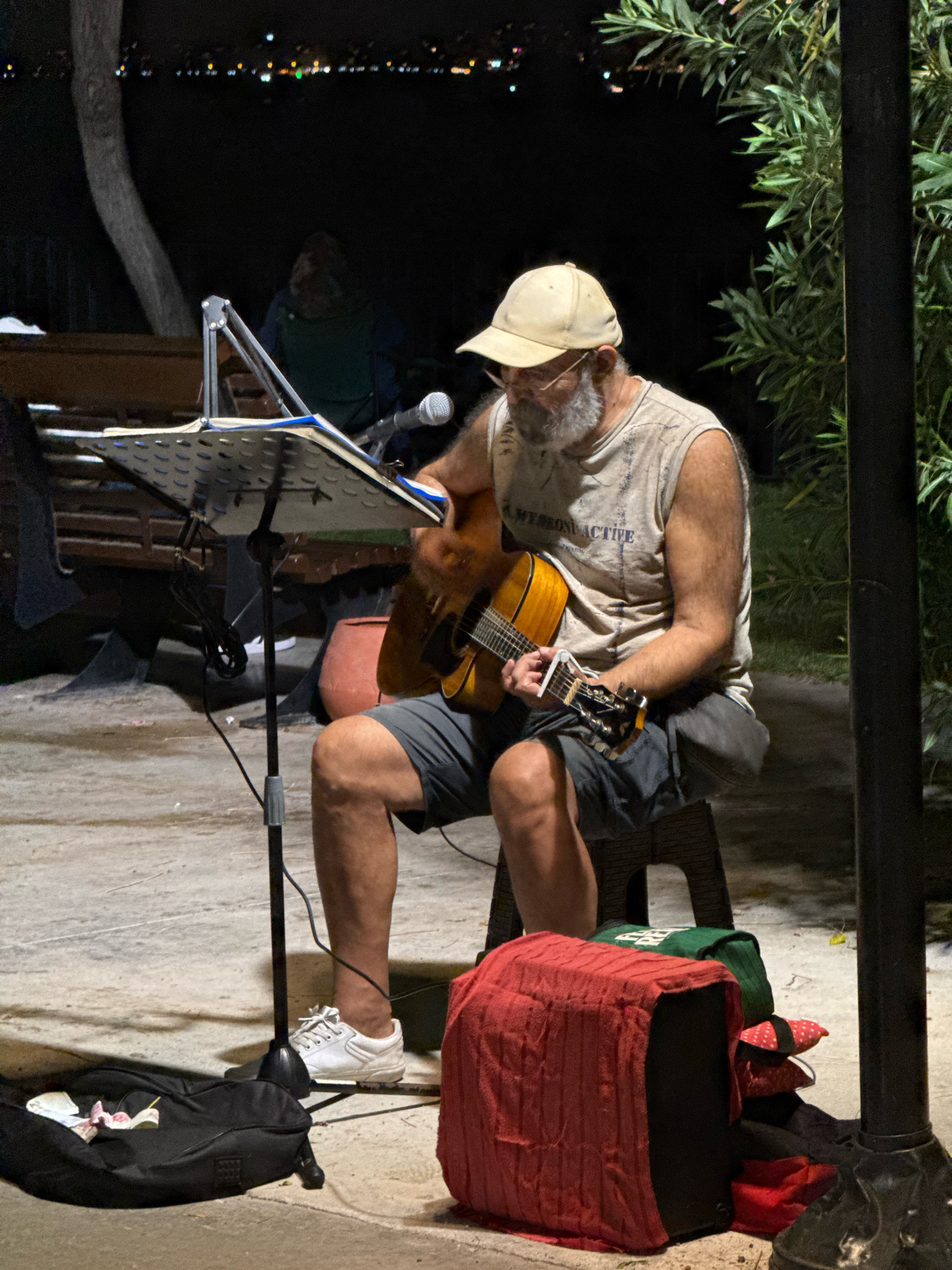 An elderly man with a grey beard and glasses is playing a guitar outdoors at night. He is wearing a light-colored sleeveless shirt, grey shorts, and white sneakers. The man is seated on a stool, and a music stand with papers is positioned in front of him. A microphone is attached to the music stand. To his right, a dark pole is visible. In the foreground, there is a black bag with some papers and a red-covered object next to it. In the background, there is a blurred figure seated on a bench and some trees with foliage. The scene appears to be in a public area with paved ground. The distant background shows blurry lights, suggesting an urban environment. The overall lighting suggests nighttime. Given the context provided, this scene is located in Konyaaltı, Turkey.