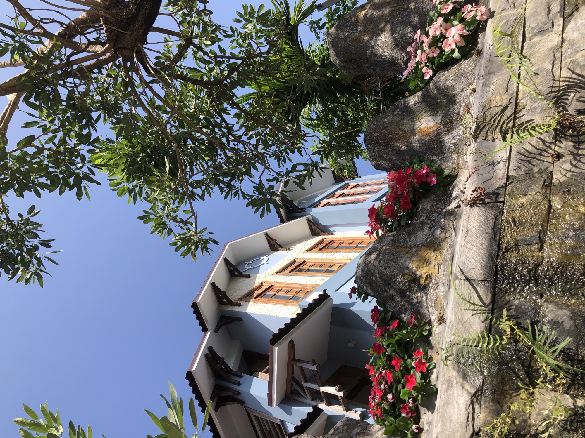 A low-angle, full shot captures the side of a blue, two-story house with a brown tiled roof in a tropical environment. The house has wooden-framed windows and ornate, triangular roof supports. In front of the house, large, rough gray rocks are covered in lush greenery, including vibrant red and pink flowers and ferns. A small waterfall cascades down the rocks, glistening in the sunlight. The sky above is a clear, bright blue, and the branches of a large tree with green leaves frame the top of the image. This scene is located at the Furama Villas in Đà Nẵng, Vietnam.