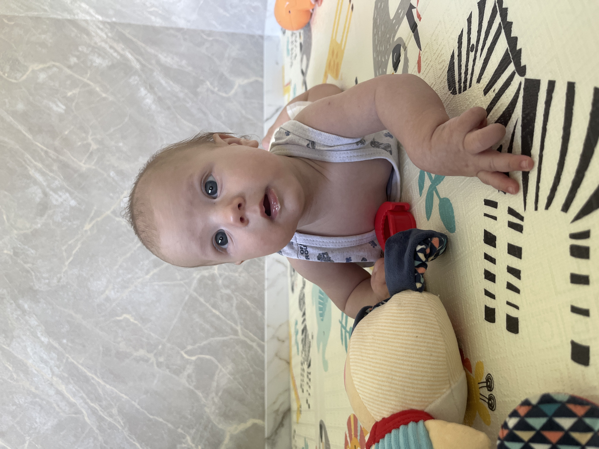 A baby is lying on its stomach on a playmat. The baby has light brown hair and blue eyes and is wearing a white tank top and a diaper. The baby is looking at the camera with its mouth slightly open. To the left of the baby is a stuffed toy. To the right of the baby is an orange toy. The background is a wall with a marble pattern. The scene appears to be indoors.