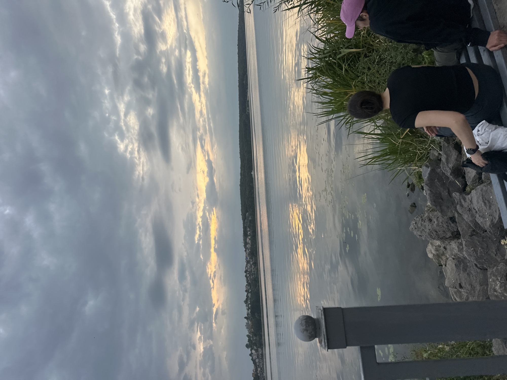 This image depicts a tranquil scene by a body of water, likely Ternopil Lake in Ternopil, Ukraine, given the presence of a distant treeline with buildings suggesting an urban or suburban edge. Two individuals are seated on a bench facing the water. The person on the left, seen from behind, wears a black t-shirt and dark pants. Their hair is tied back. They are looking out at the water. To their right, another person, also seen from behind, is wearing a black hooded sweatshirt and a pink baseball cap. They are also facing the water. The bench is situated on a rocky shore with tall green reeds growing nearby. In the foreground, a dark metal railing with a round finial is visible on the left. The water is calm, reflecting the cloudy sky and a band of golden light peeking through the clouds on the horizon. The overall atmosphere is one of quiet contemplation or relaxation by the water.