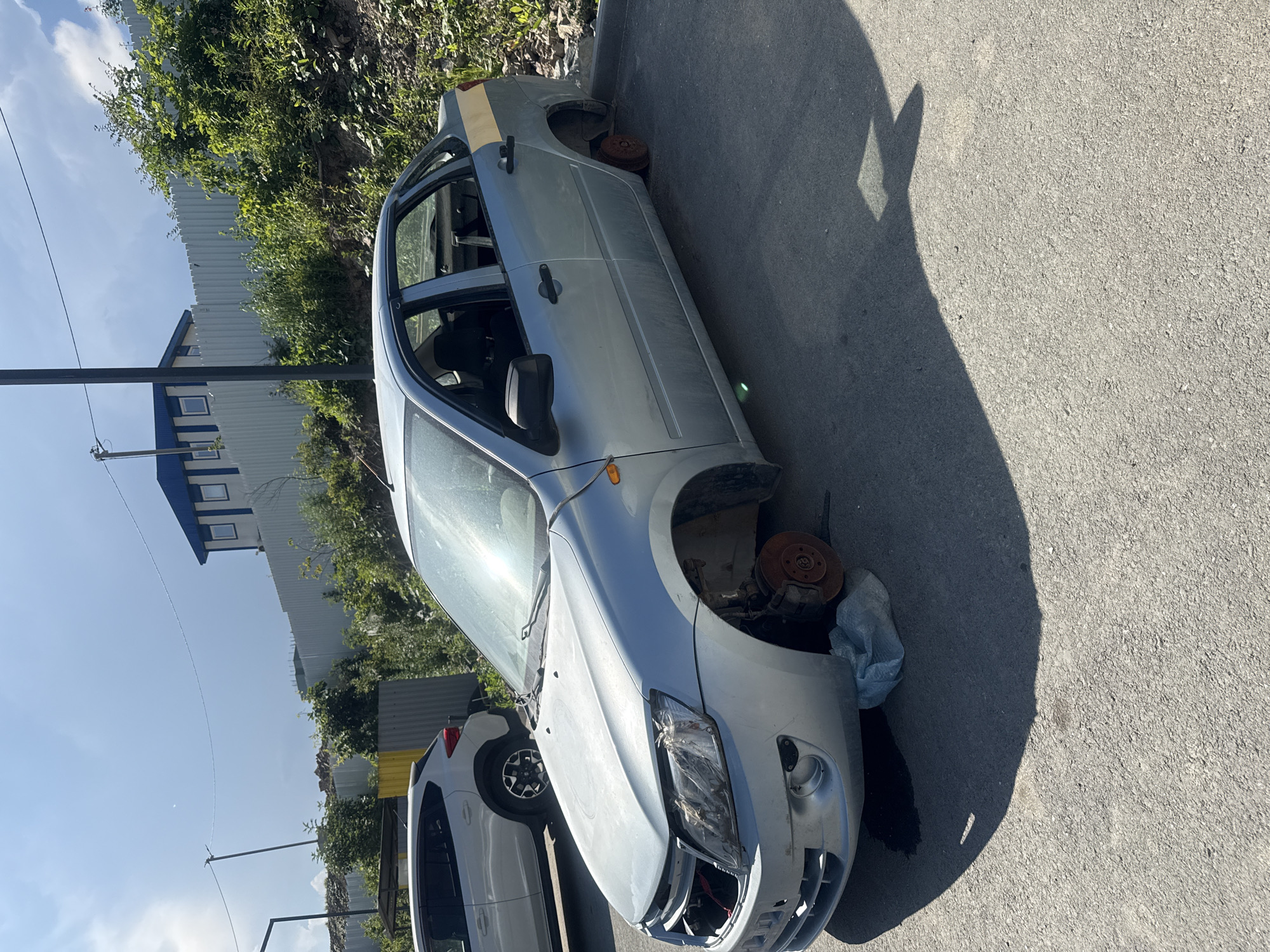 A silver sedan, appearing to be a Lada Granta, is parked on an asphalt surface. The car is missing its wheels, and its front bumper and headlights are damaged. A blue plastic bag is visible near the front passenger-side wheel well. The scene is outdoors, under a clear, bright sky, suggesting a sunny day. The sun casts a long shadow from the car onto the asphalt.
In the background, there is a tall, light-colored corrugated metal fence and lush green foliage. Beyond the fence, a building with a blue roof and white facade can be seen. Another white car is partially visible in the lower left quadrant of the image, parked behind the sedan.
The overall impression is that of a vehicle that has been stripped for parts or is in a state of disrepair. There is no visible text in the image. The context provided indicates the location is Vladivostok, Russia. The time of day appears to be midday or early afternoon, given the strong sunlight and the length of the shadow. The weather seems to be warm and clear.