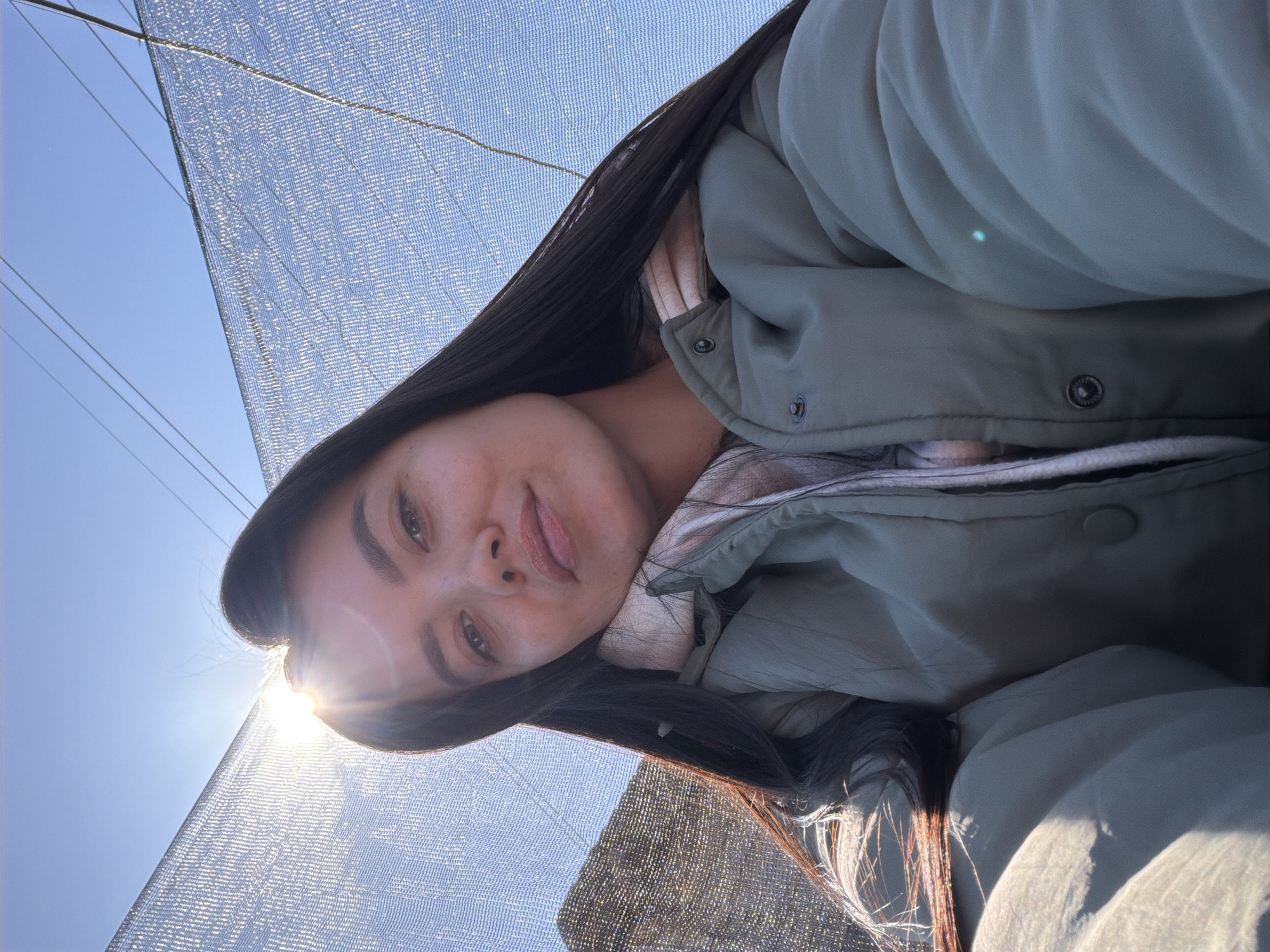 A person is visible in the image. They appear to be wearing a green jacket over a lighter-colored garment, possibly a hoodie or sweater. Long, dark hair is visible, partially obscuring their face. The individual is positioned beneath a semi-transparent netting, possibly a shade cloth, with a blue sky and sunlight visible in the background. Based on the scene and the context provided, the location is likely Chaikovsky, Russia.