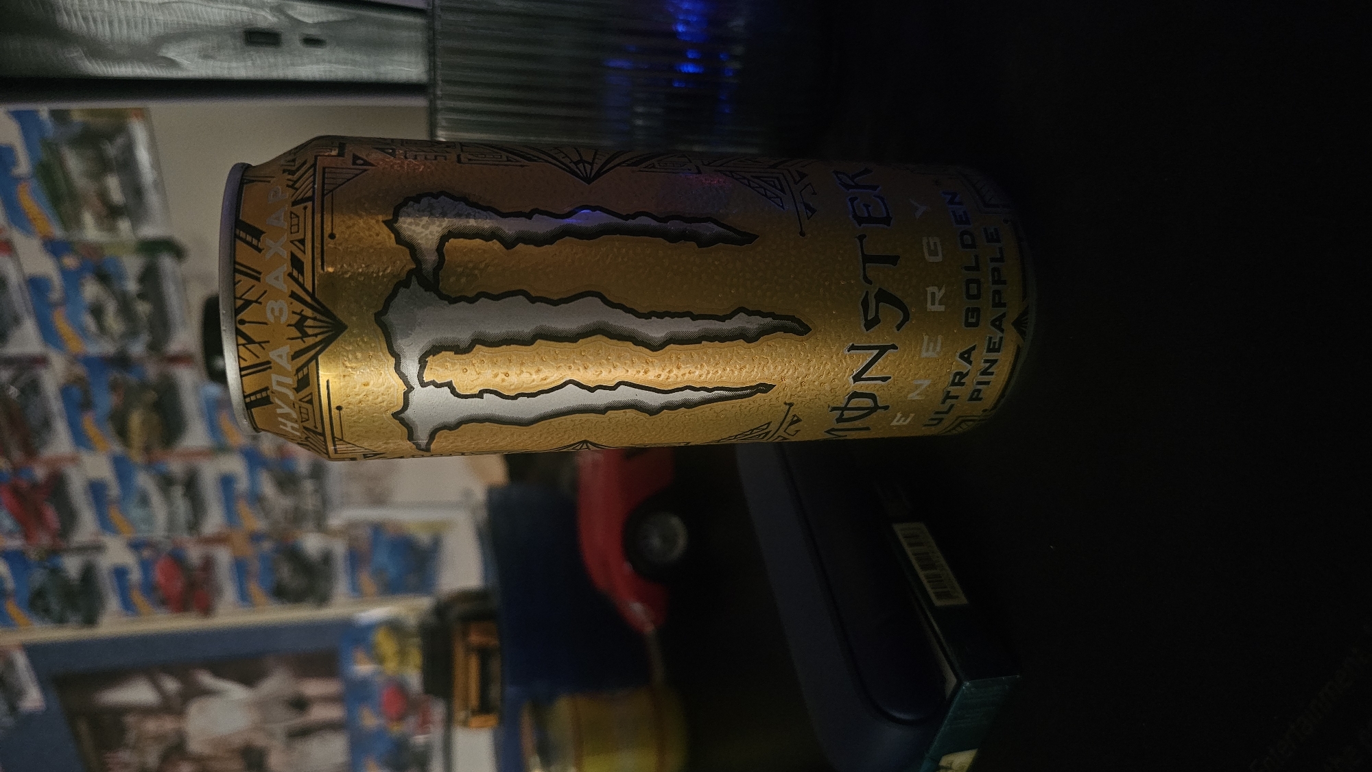 A horizontally oriented, close-up shot features a Monster Energy "Ultra Golden Pineapple" drink can. The can is mostly gold with silver accents and black text displaying "MONSTER ENERGY ULTRA GOLDEN PINEAPPLE" along with the iconic Monster claw logo. Condensation droplets are visible on the can's surface. The can is surrounded by clutter, and is resting on a dark surface. To the left of the can, a blurred background shows a shelf of Hot Wheels cars. Parts of an electronic device are visible near the lower right of the frame.
There is no explicit information to place this scene in Varna, Bulgaria.