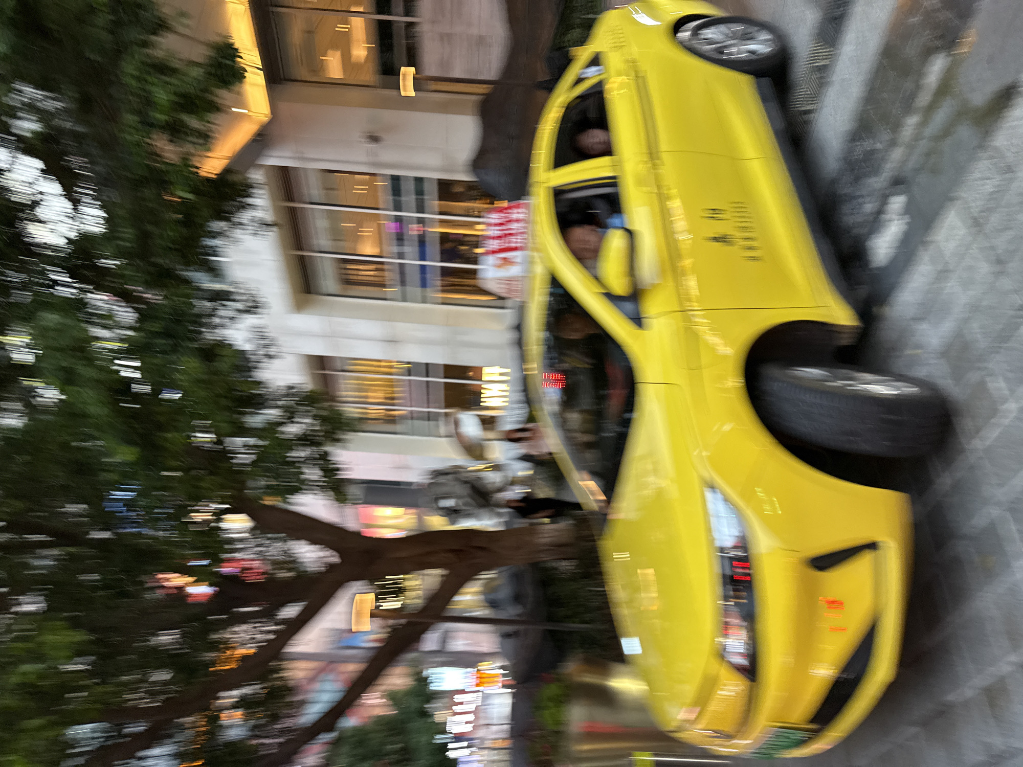 A bright yellow taxi is captured in motion on a wet street in Yuzhong District, China. The taxi has passengers inside and is illuminated by streetlights and building lights. The scene is blurred due to the movement of the vehicle and possibly a low shutter speed, creating streaks of light and color. A large tree with dark foliage is on the left side of the image, and a modern building with glass windows and illuminated interiors is visible in the background. The ground appears to be paved with stone or tile and reflects the surrounding lights.