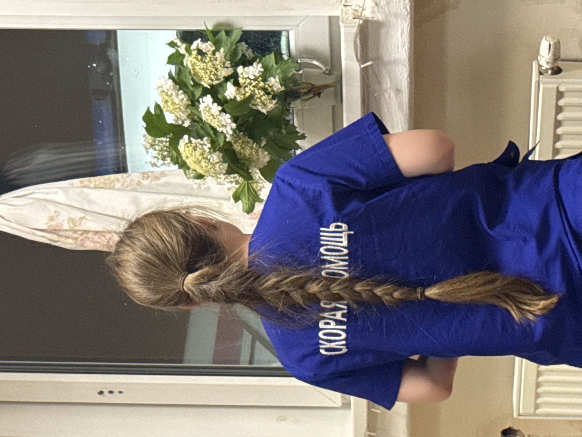 A woman with a blonde braid stands in front of a window with white frames and a radiator. The woman is wearing a blue t-shirt with white text that reads "СКОРАЯ ПОМОЩЬ" ("Ambulance"). There is a vase of white flowers on the windowsill. The location is potentially the city of Chaikovsky, Russia.