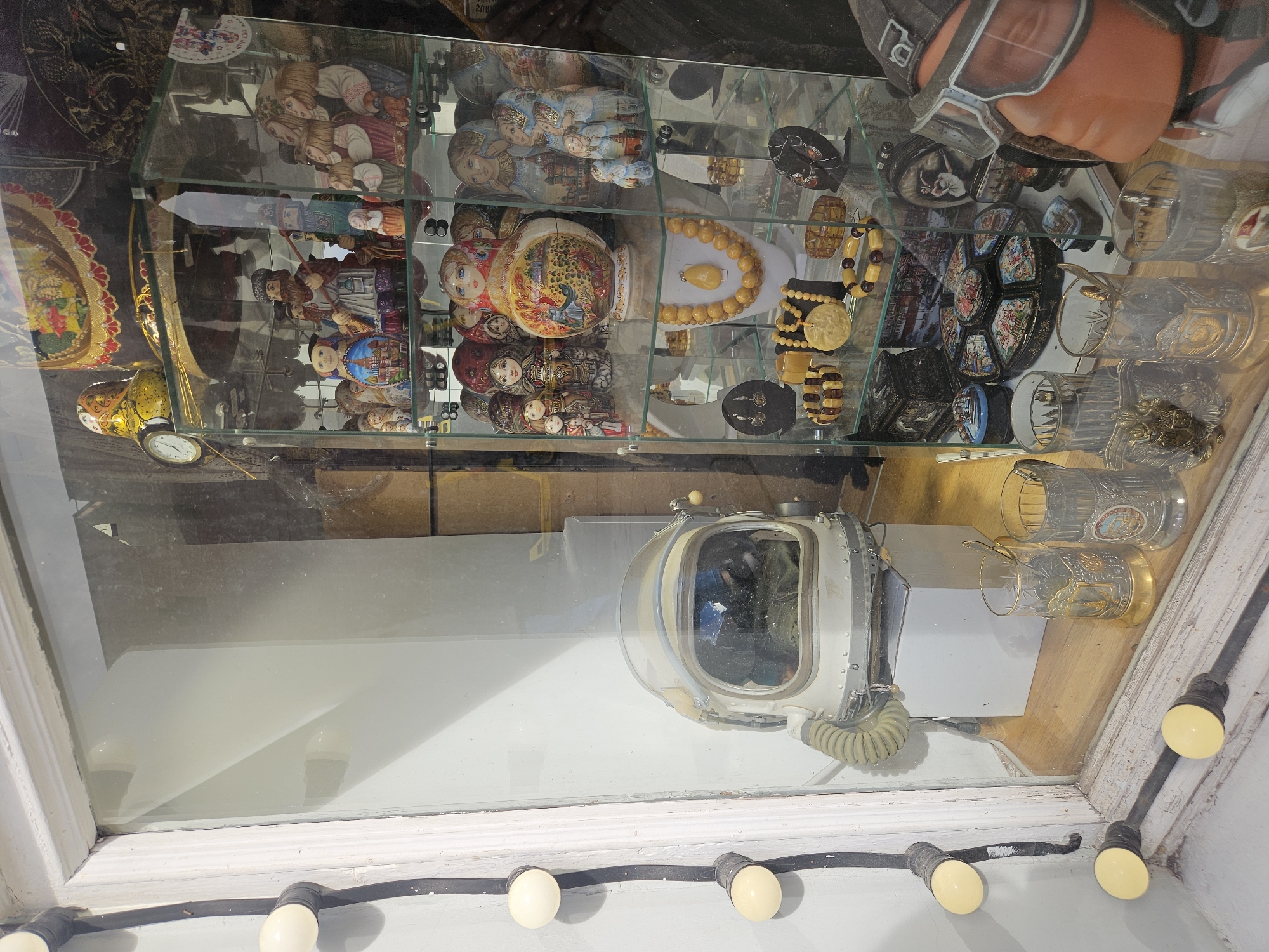 The photograph shows a display window containing various artifacts. At the top of the window, there is a glass shelf filled with items, including Russian nesting dolls (Matryoshka dolls), jewelry, and decorative plates. At the bottom of the window, there is a vintage-style diving helmet with a clear dome. The photograph is taken from outside the window, showing the artifacts inside. The location is the city of Unknown, Unknown.
